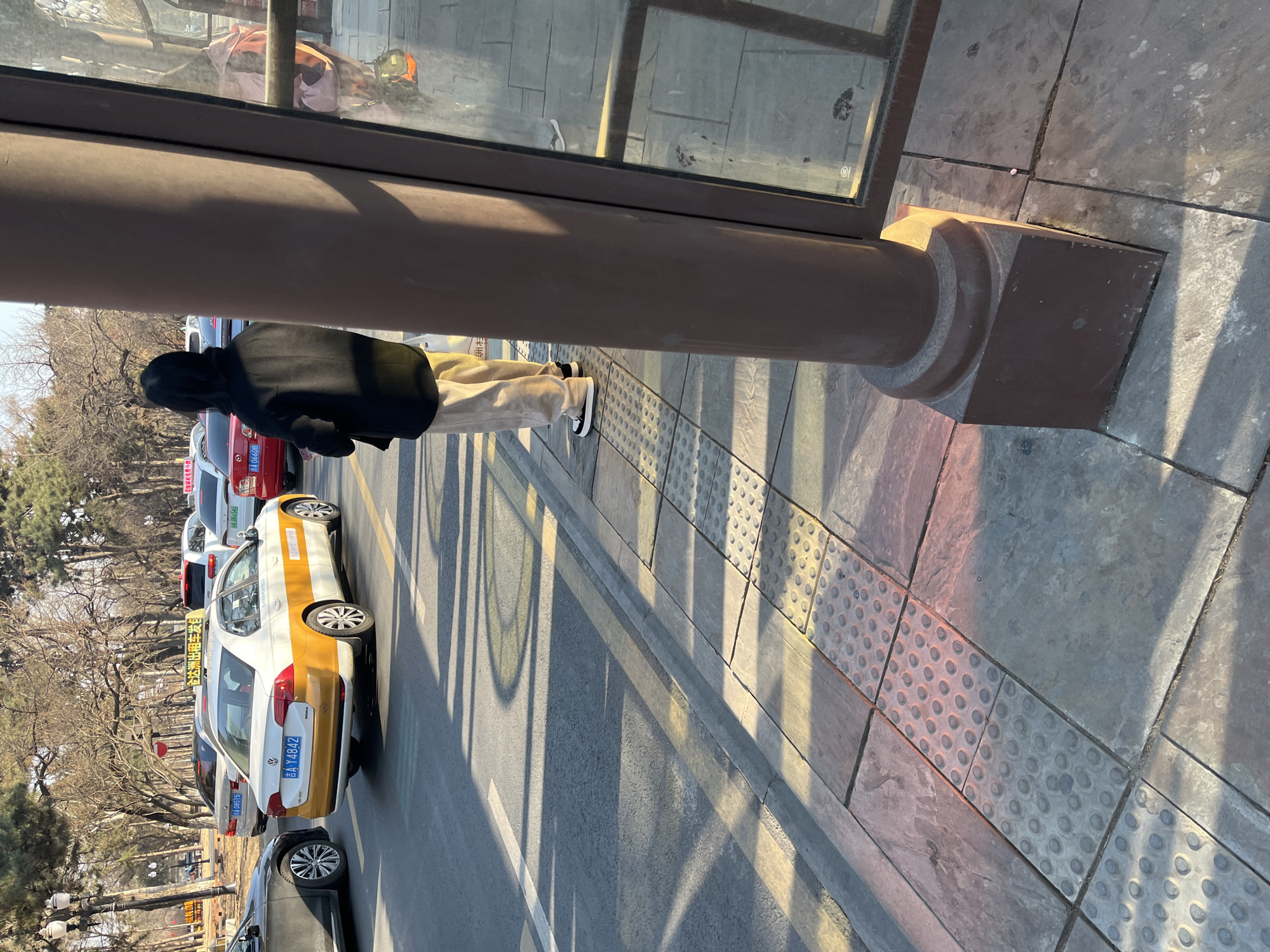 The image shows a person standing at a bus stop in Changchun City, China. The person is wearing a dark jacket and light-colored pants, and is facing away from the camera. The bus stop has a brown pillar and glass panels. In the background, there is a street with several cars parked along the side. A taxi is visible, and its license plate reads "吉A-Y4842". The text on the taxi reads "金达洲出租车发". The other license plates visible read "BA 08515", "BA 061545", and "BA 06608". There are trees in the background, suggesting the location is near a park or tree-lined street. The sidewalk has tactile paving near the curb.