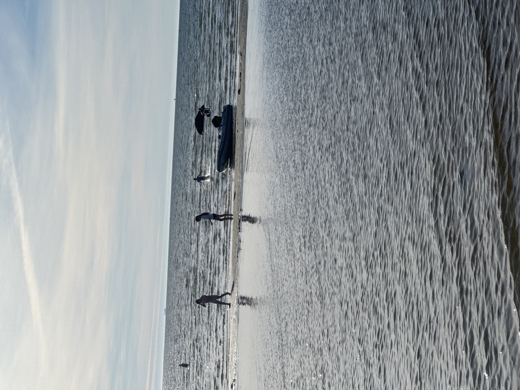 The image depicts a calm sea with a sandy beach in the foreground. The sky is partly cloudy with visible streaks of white clouds. Several individuals are visible in the water and on the shoreline. In the foreground, a shallow body of water reflects the sky and ripples across its surface. Further back, on the sandbar and in the shallow sea, three figures are walking or standing. One figure, on the left, is silhouetted and appears to be walking into the water. Two other figures are closer to the right, one of whom is also silhouetted. In the mid-ground, an inflatable boat is partially beached on the sandbar, with a person standing near it in the water. There are also two dark, rounded objects in the water beyond the boat, which could be buoys or rocks, with at least one person interacting with them. In the distance, a large ship is visible on the horizon, as are a few small, indistinct objects that could be buoys or other vessels. The overall scene is one of a leisurely day at the beach. Given the presence of a large ship on the horizon and the general appearance of the sea, it is plausible that this scene is located on the coast of Tallinn, Estonia, which has a significant port and coastline along the Baltic Sea.