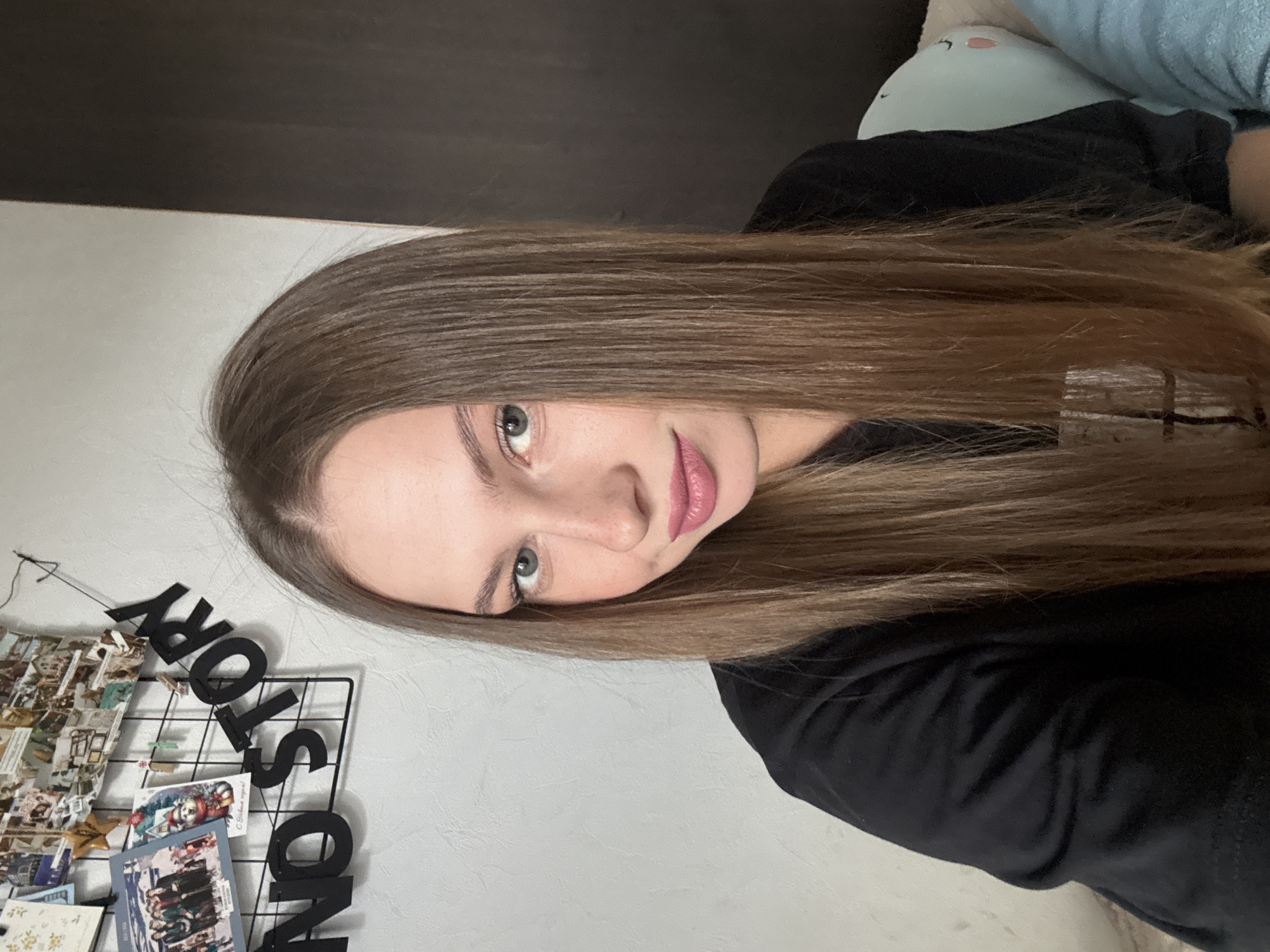 A young woman with long, straight brown hair and blue eyes is captured in a close-up, head-and-shoulders selfie. She is wearing a black t-shirt and is looking directly at the camera with a slight smile. Her makeup includes pink lipstick and subtle eyeliner. Behind her, a wall is decorated with a grid filled with photos and a banner that reads "NO STORY". The lighting suggests an indoor setting, possibly a bedroom, with a darker area on the right side of the frame and a hint of a pillow or stuffed animal. The overall impression is casual and personal. There is no specific indication of the city of Perm, Russia within the image itself.
