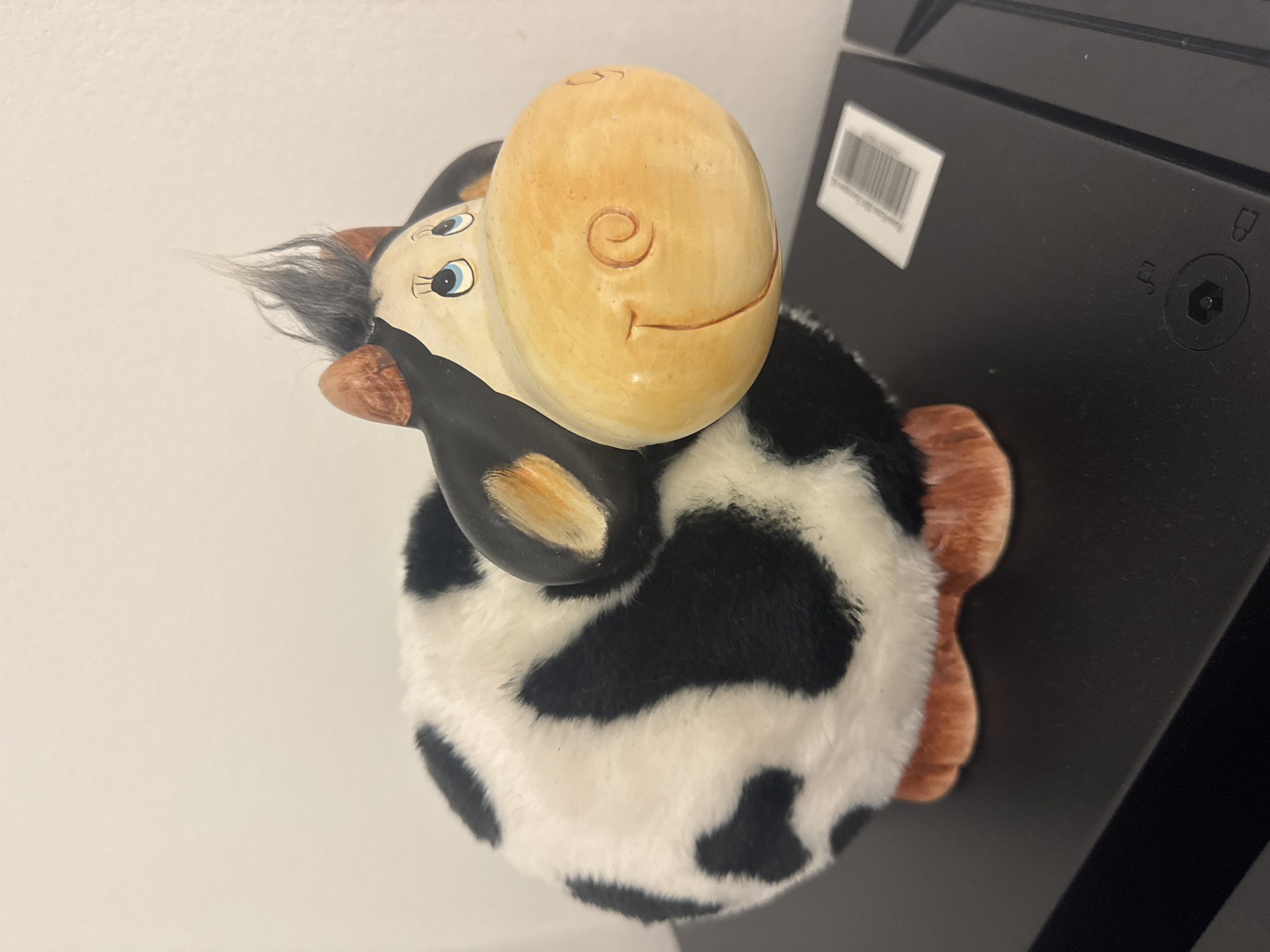 A close-up, high-angle shot shows a stuffed cow toy with a ceramic head placed on a dark, possibly black, surface. The cow has a round, cream-colored head with a painted smile and a curled horn on its cheek. Its body is covered in black and white fur, resembling a Holstein cow. The toy has brown ears and a tuft of gray hair on its forehead. A barcode sticker is visible on the black surface behind the cow. The location is unspecified; no elements indicate it being in Sofia, Bulgaria.