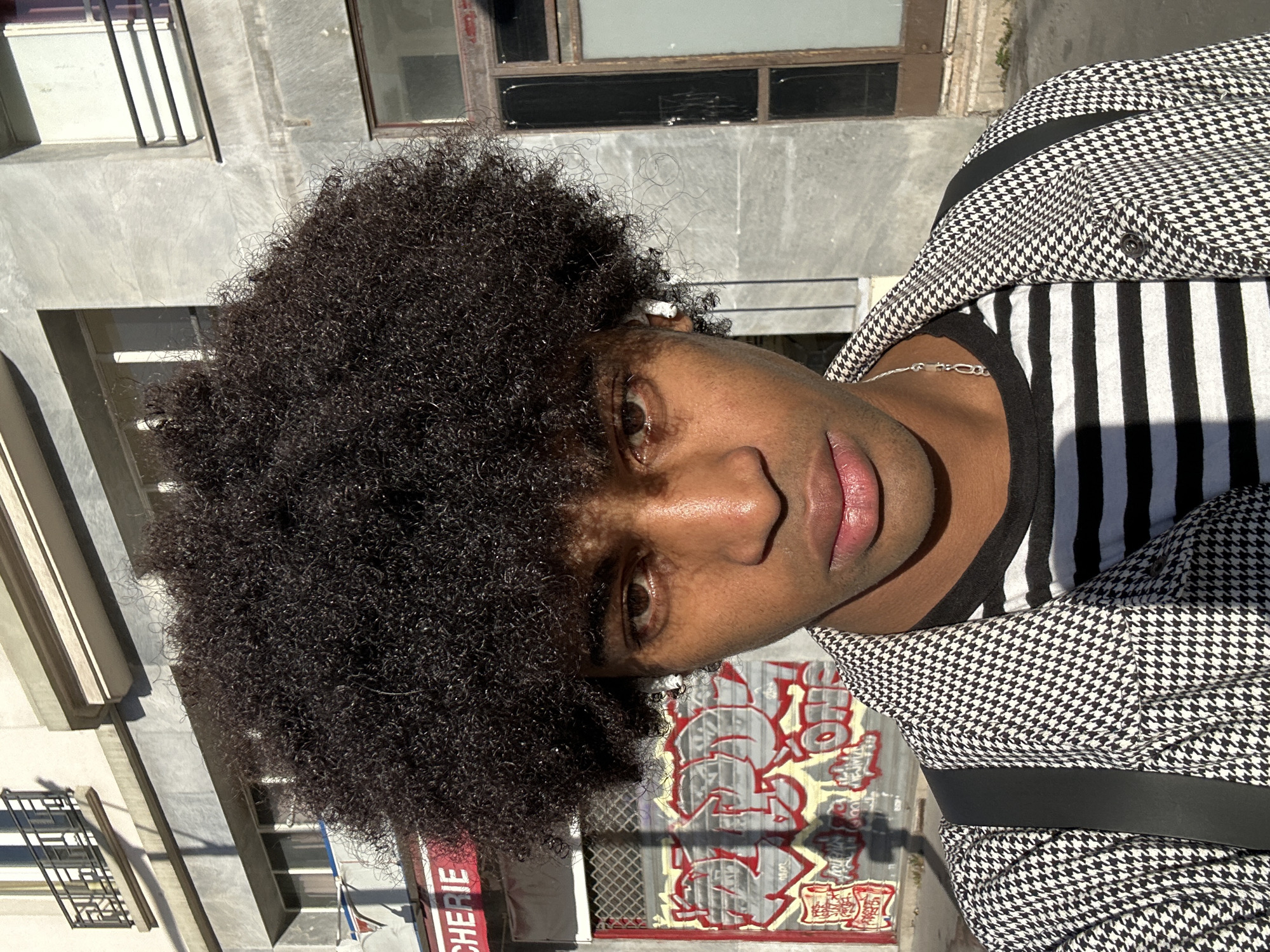 A medium shot depicts a person with dark curly hair, wearing a black and white striped shirt underneath a black and white houndstooth jacket. A silver chain necklace is visible around the person's neck.
The background includes an urban scene with a building to the left. Fencing and graffiti are visible in the lower portion of the frame. A sign with the partial word "CHERIE" in red letters is also visible.
Based on the architectural style and signage, the image is likely taken in Paris, France.