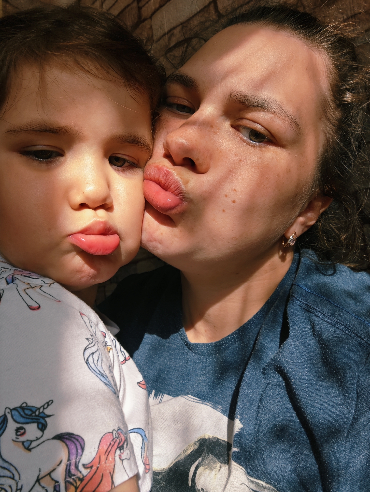 A close-up indoor shot shows a woman with freckles kissing a young child on the cheek. Both are pouting with exaggerated lip expressions. The child wears a white shirt with unicorn print. The woman wears a denim-like blue shirt and a simple earring. The lighting suggests a sunny day. The background is a wall with a rough texture. There is no evidence to suggest a location in the city of Уральское, Russia.