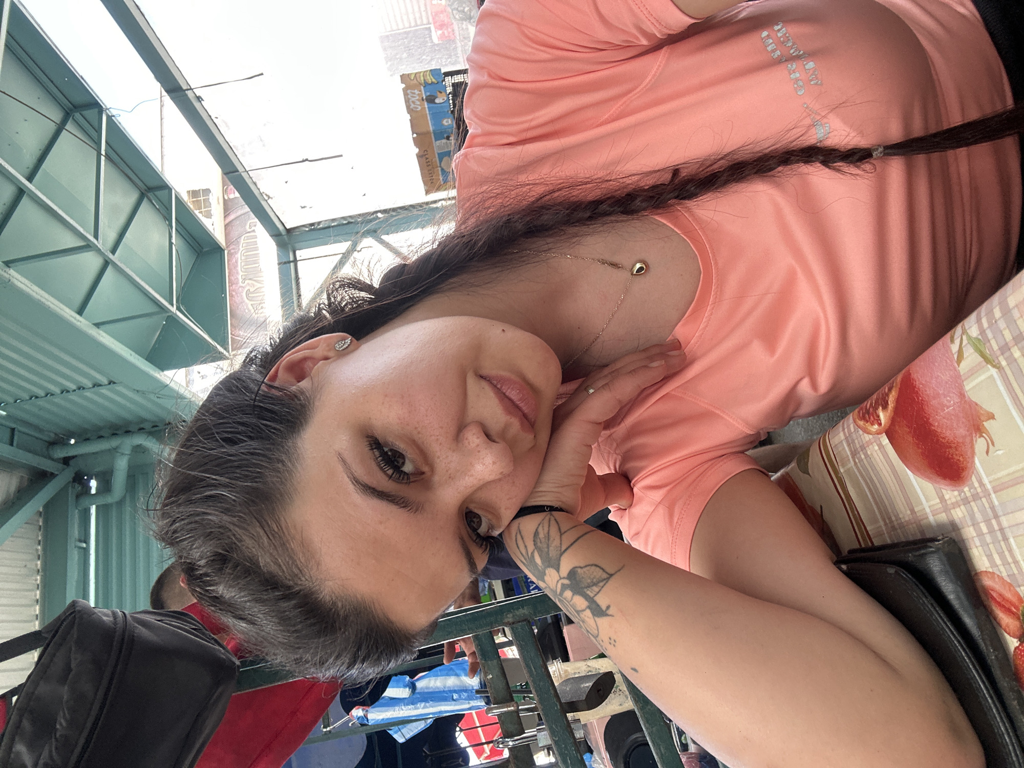 A young woman with dark hair in a braid and freckles across her nose is captured in a selfie. She is wearing a coral-colored t-shirt with subtle silver lettering on the right side. Her left arm rests on a table covered with a checkered tablecloth featuring images of fruit. A black wallet is placed beneath her arm. She has a tattoo on her left forearm. The background appears to be an outdoor market or food court with a green metal roof structure overhead, indicating a location in Sliven, Bulgaria, likely at the market or in the vicinity of the city's bus station, which has similar architectural features. The overall lighting suggests daytime.