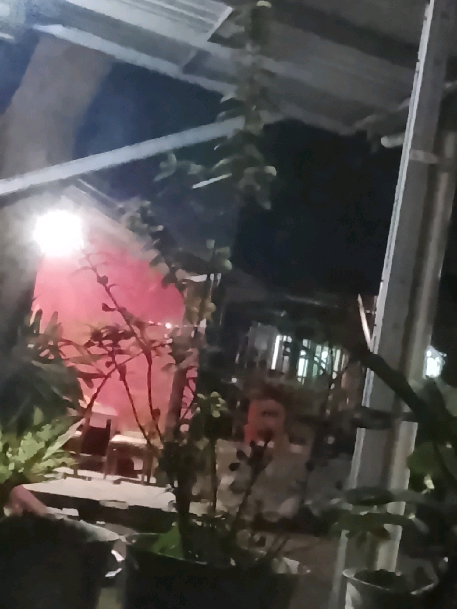 The image depicts an outdoor scene at night, possibly at a cafe or restaurant in Ujungrusi, Indonesia. A bright light illuminates a pink-colored structure or wall in the background, with a tall, thin plant in the foreground obscuring some of the view. To the left, a table is visible, with another object that could be a chair behind it. The overall impression is of a dimly lit, outdoor dining or gathering space.
