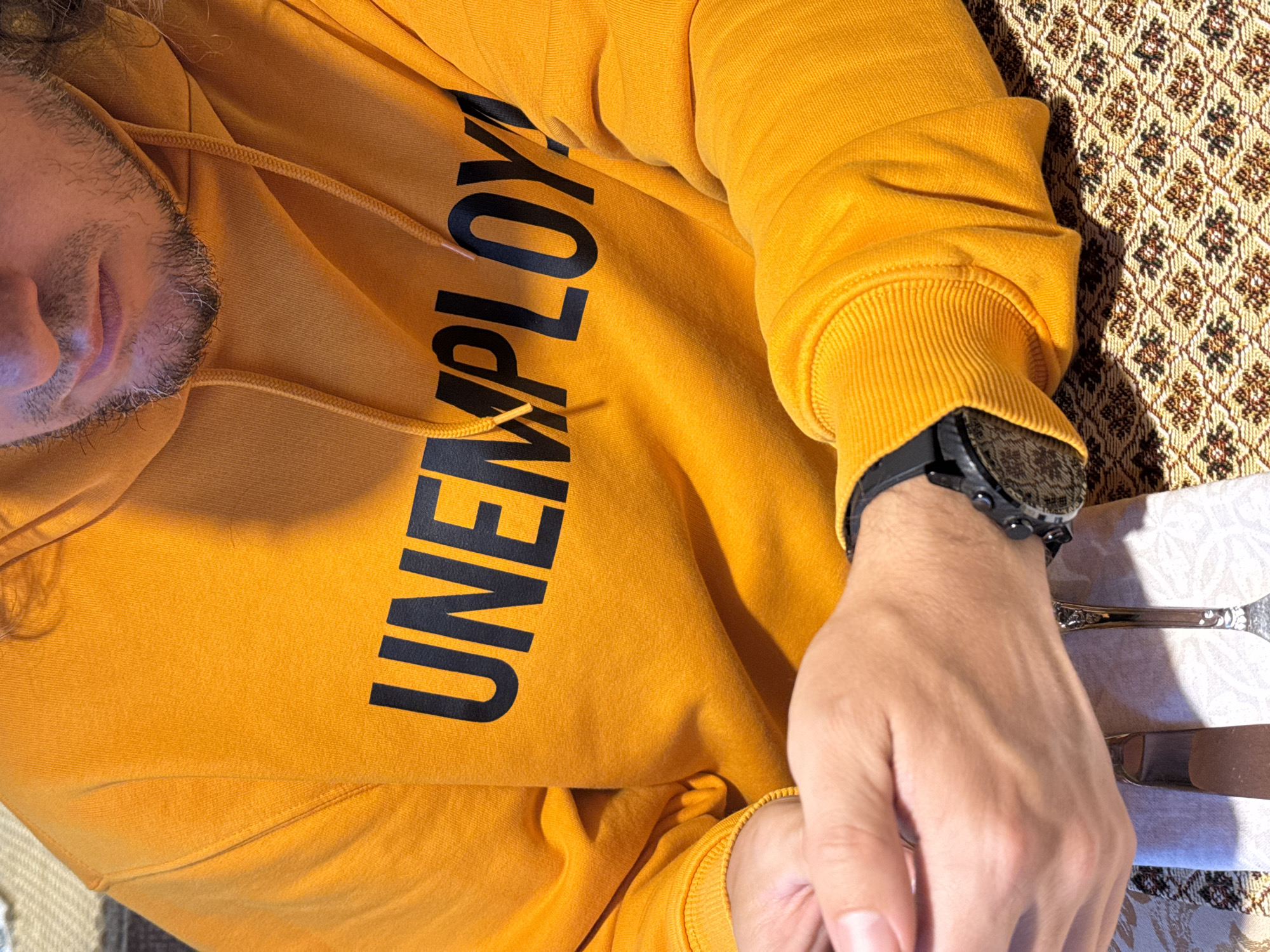 The image shows a person wearing a yellow hoodie with the word "UNEMPLOY" printed on it. The person's face is partially visible, showing a beard. They are lying on a surface with a patterned tablecloth. A watch is on the person's left wrist. Cutlery is placed on a white napkin next to the person's hand. The location is not identifiable as Ketoshi, Russia, or any other specific location.