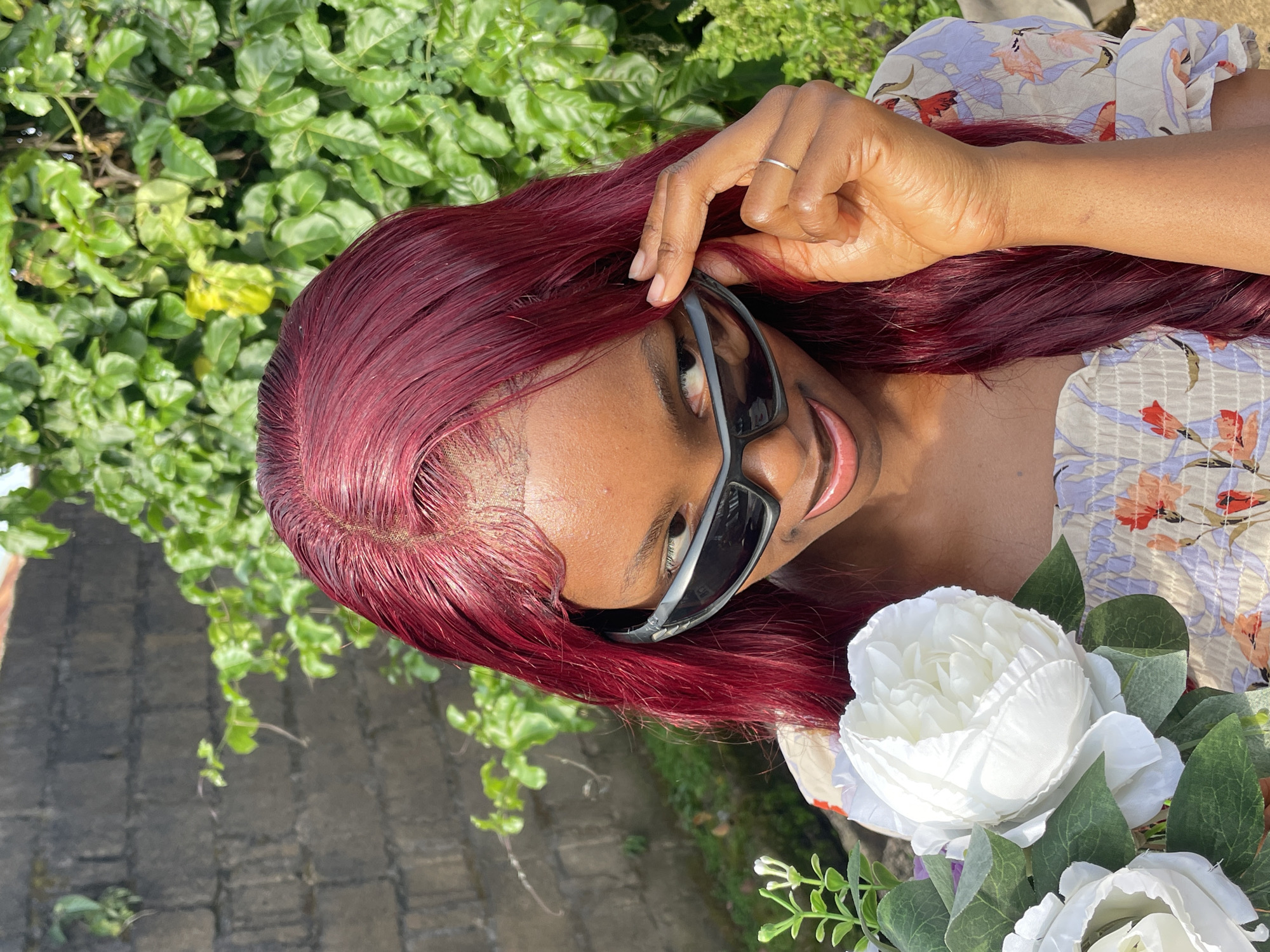 A close-up, horizontally oriented, eye-level shot shows a person with dark skin and long, dark red hair. The person is wearing dark sunglasses and a light-colored, floral-patterned top. They are touching their hair and sunglasses with their right hand, which is adorned with a silver ring. The person's mouth is slightly open, and they appear to be smiling. To the lower right of the frame, a bouquet of white flowers with green leaves is visible. The background consists of lush green foliage and a stone wall, suggesting an outdoor setting in a sunny environment. The overall mood is relaxed and cheerful. There is no visible text in the image. The location context is Jos, Nigeria.