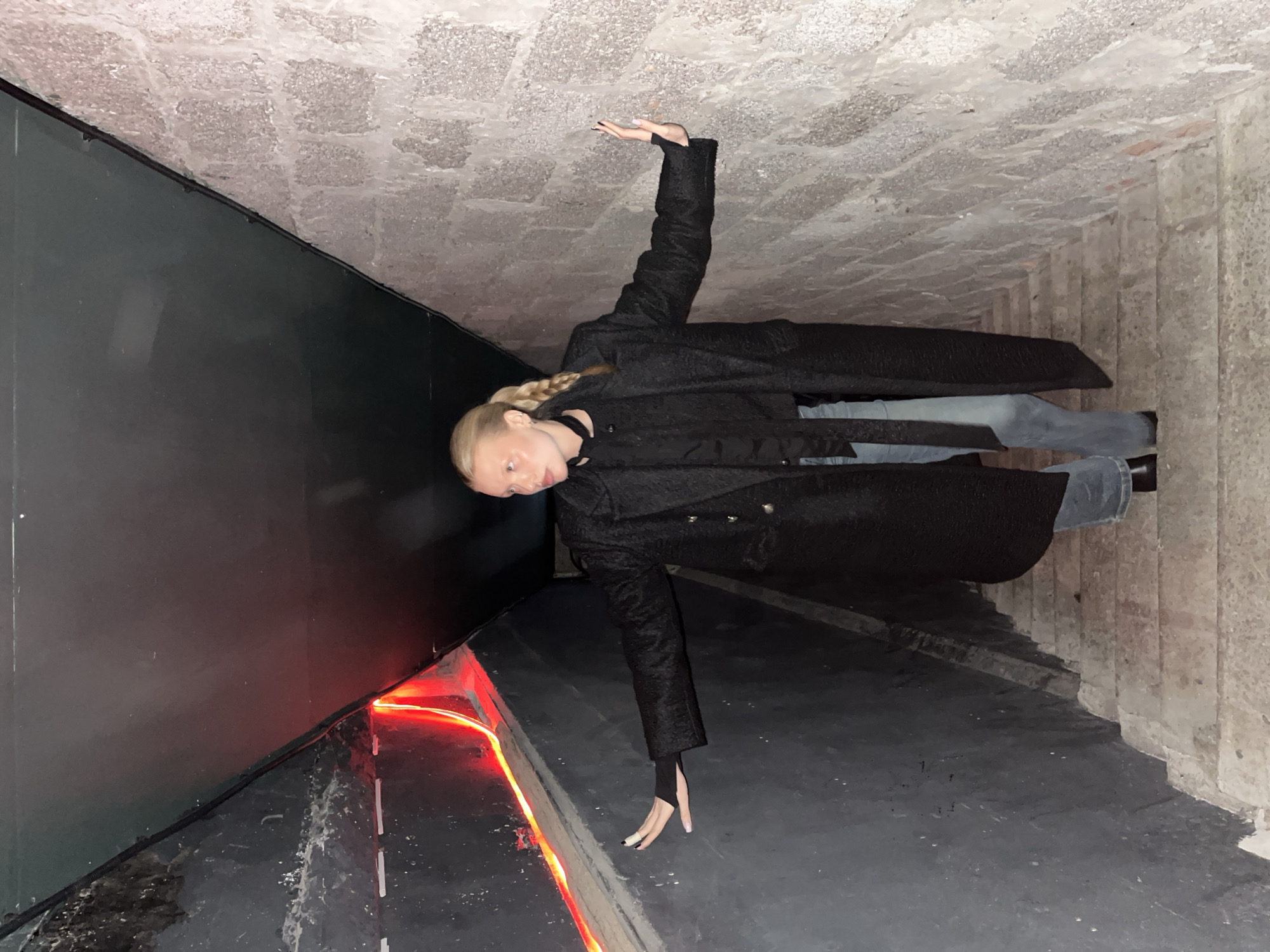 The image depicts a blonde woman in a black coat and blue jeans standing in what appears to be an underground tunnel or passageway in the city of Chaikovsky, Russia. One arm is raised overhead, appearing to touch the stone brick ceiling. The other arm extends downward, her hand near a strip of red light. The passageway has gray concrete walls and floor, and the ceiling is made of rough stone bricks. The woman is facing forward, slightly tilted.