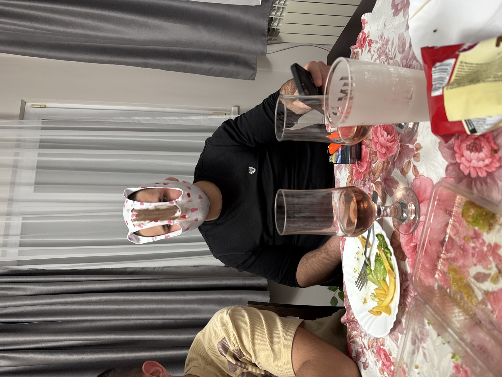 A tilted medium indoor shot shows a man wearing a black shirt and a head covering with pink hearts. He is holding a black smartphone and a glass. The scene takes place around a table covered with a floral patterned tablecloth. There are various containers of food, a plate with remaining salad and french fries, another glass, and a pack of cigarettes on the table. Another person wearing a brown and cream patterned shirt can be seen seated. The setting appears to be a dining area, possibly in a home in Topoli, Bulgaria. The exact location within Topoli is not discernible from the image. A window with curtains is visible in the background.