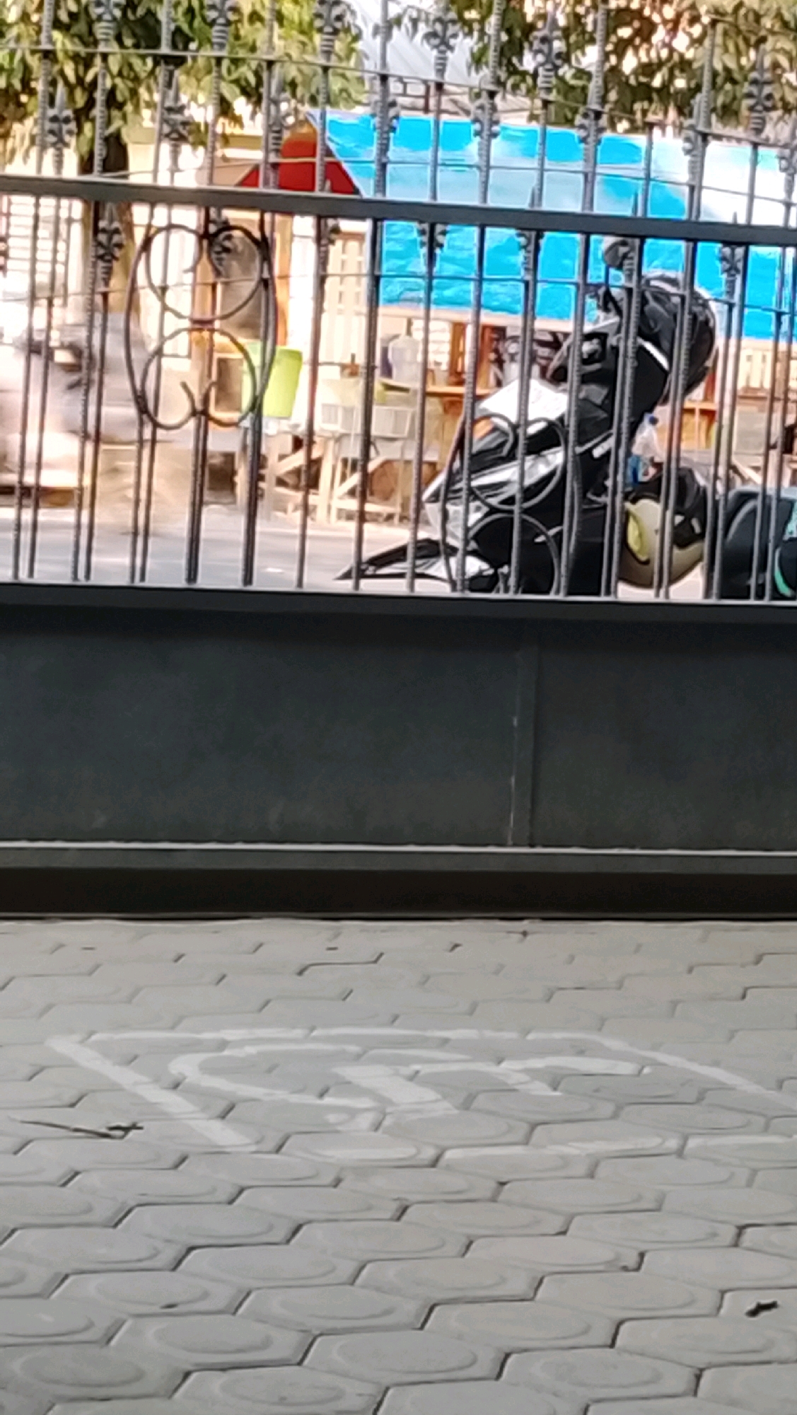 The image shows a portion of a black metal fence with decorative scrollwork. Behind the fence, a motorcycle is visible, parked under a blue tarp or awning. The motorcycle is black and white with some reflective detailing. In the background, through the fence, there are glimpses of buildings and trees. The foreground shows a paved area with hexagonal tiles, marked with a white symbol that appears to be a parking or accessibility sign. The scene is outdoors, with natural light suggesting it is daytime. The location is identified as Slawi, Indonesia.