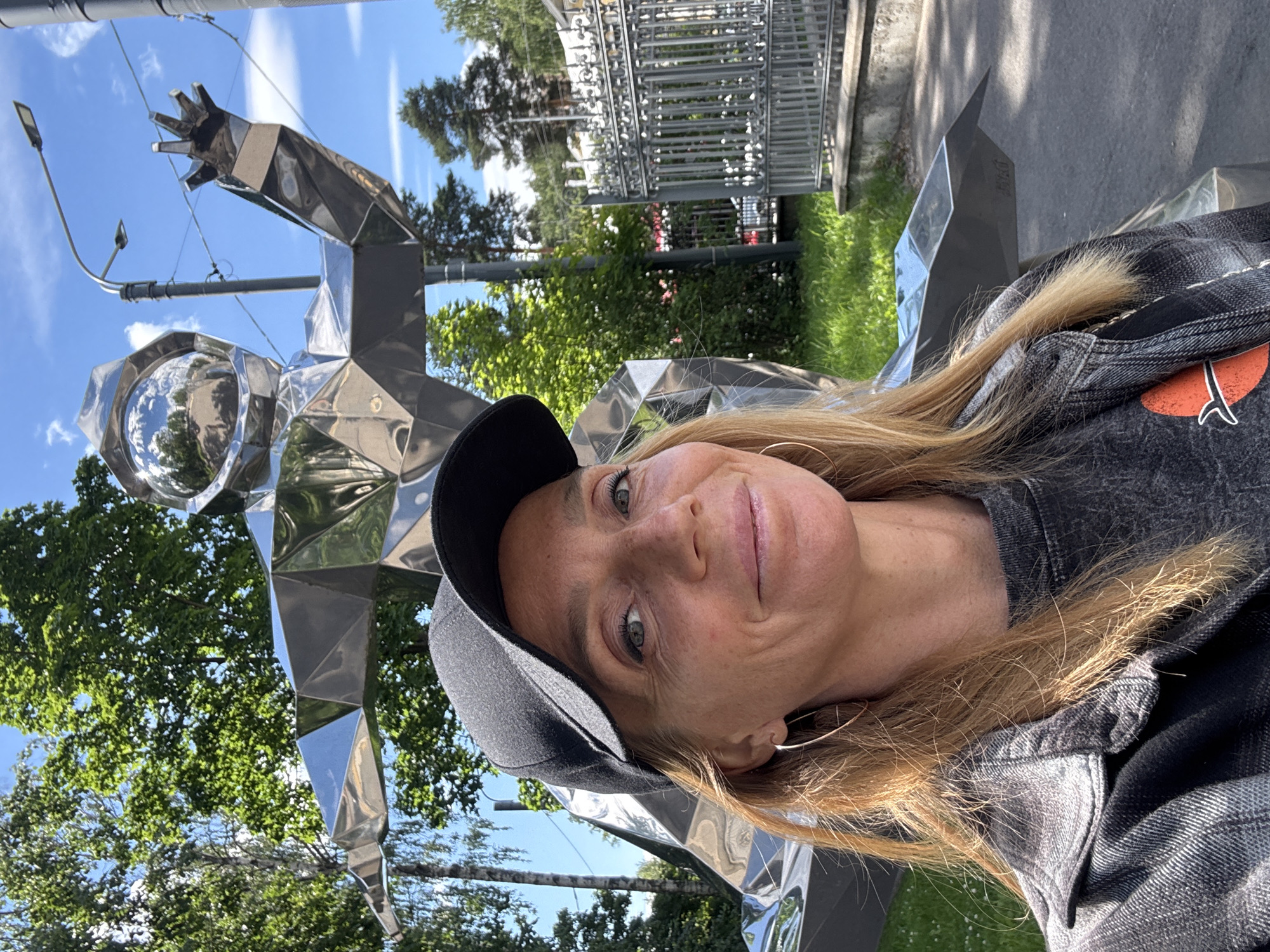 A woman with long blonde hair, wearing a black baseball cap and a grey denim jacket, smiles into the camera. She is outdoors with a shiny, angular metal sculpture in the background. The sculpture appears to be abstract and futuristic, with reflective surfaces that mirror the surroundings. Behind the sculpture and the woman, lush green trees and a bright blue sky with scattered clouds are visible. In the distance, a metal fence and a building are partially visible. The lighting suggests it is daytime, likely a sunny day, given the brightness and clear sky. The woman's expression is cheerful and relaxed, indicating a pleasant outdoor experience. The location is identified as Sinyavino, Russia. There is no visible text in the image.
