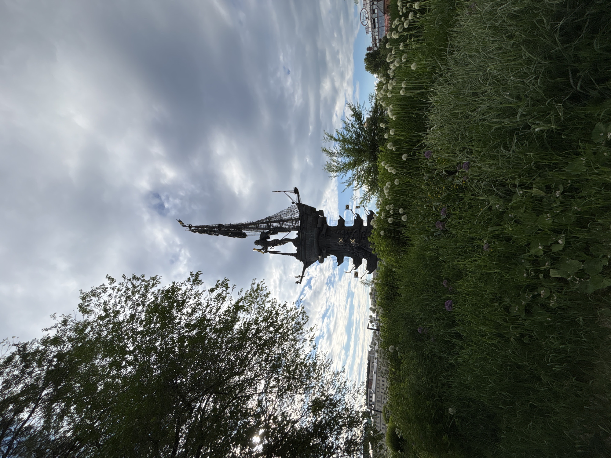 The image shows the Peter the Great Statue, in Moscow, Russia. The statue is a bronze sculpture depicting Peter the Great standing on a ship. The statue is located on an artificial island in the Moscow River. In the foreground, there are trees and tall grass. The sky is cloudy.