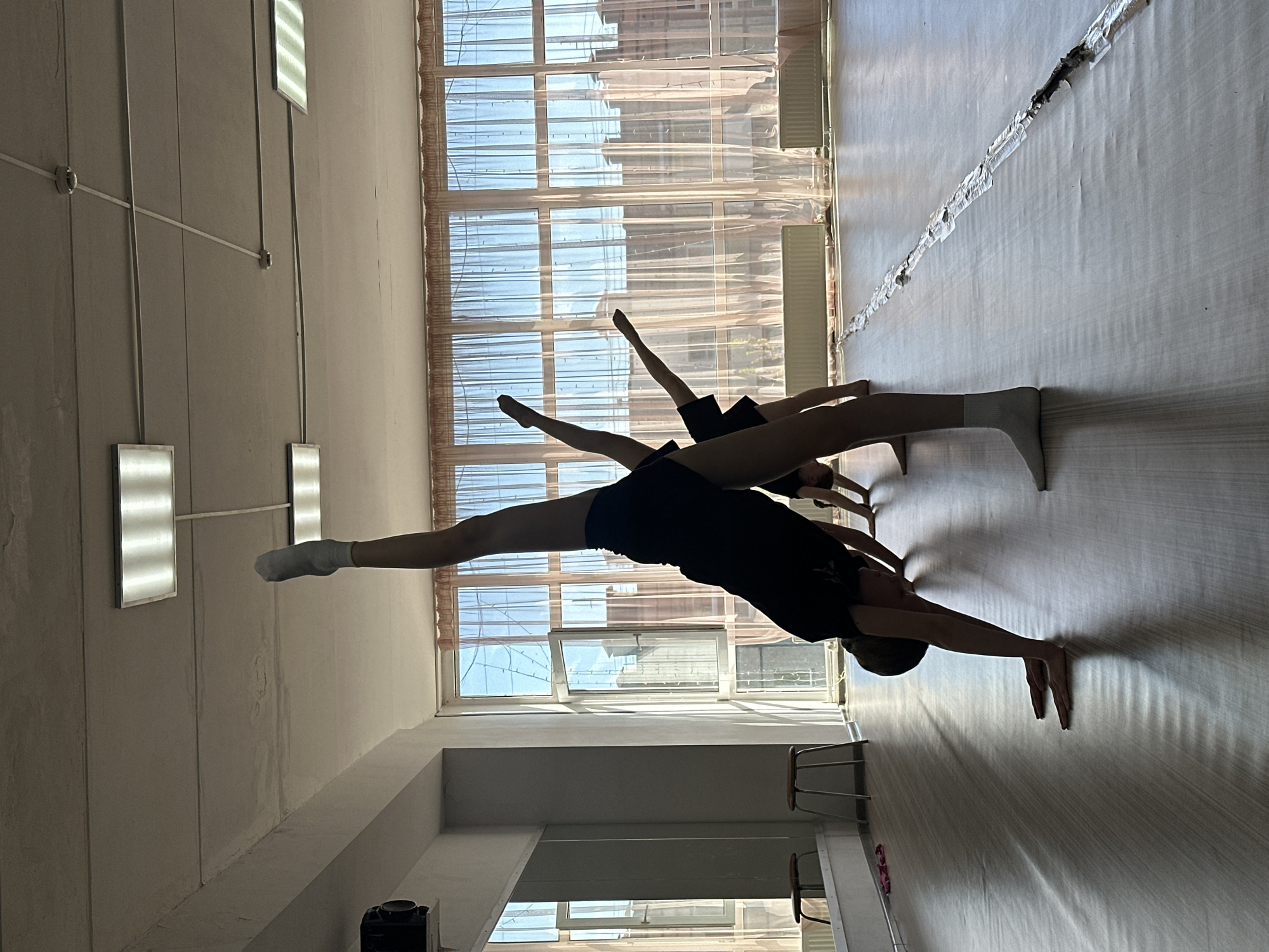 The image shows two individuals performing stretching exercises in an indoor space. The primary figure is doing a split, with one leg extended towards a large window and the other towards the opposite wall. This individual is wearing dark-colored clothing. A second person is visible behind the first, also engaged in a stretch. The room has large windows with translucent curtains. There are also light fixtures on the ceiling and chairs. The location is likely a dance or exercise studio. There is no information in the image to confirm that it is in Votkinsk, Russia.
