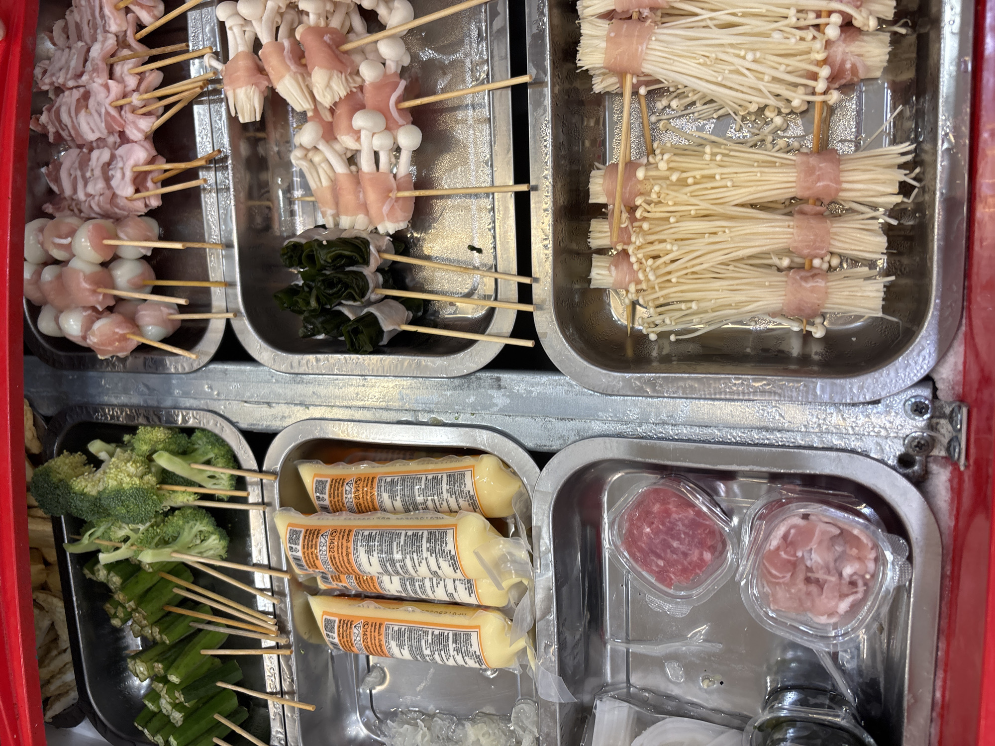 The image depicts a food cart in what appears to be a street market. The cart is filled with stainless steel trays containing various skewered food items ready for cooking or serving. The food items include: meat wrapped around enoki mushrooms, mushrooms wrapped in meat, leafy greens wrapped in meat, broccoli florets, okra, sausage sticks, and raw meat in plastic containers. The cart is red on the outside.