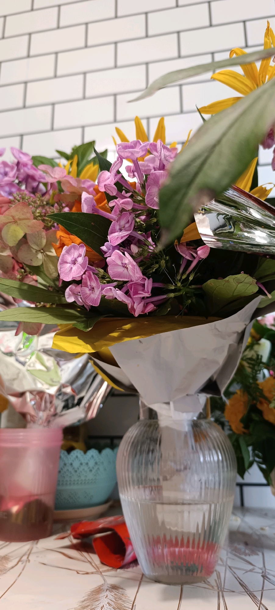 A close-up, low-angle shot captures a bouquet of flowers in a clear glass vase. The flowers are predominantly pink and yellow, with some greenery visible. The bouquet is partially wrapped in shiny silver and yellow paper. The vase sits on a textured surface with a pattern resembling wheat stalks, possibly a tablecloth. In the background, a white tiled wall is visible. To the left of the vase, a translucent pink cup and a teal decorative bowl can be seen. A red object, possibly a wrapper or small container, lies on the surface between the pink cup and the vase. The lighting is soft, creating a slightly diffused atmosphere. The scene is indoors, with no specific indicators of the city of Chaikovsky, Russia.