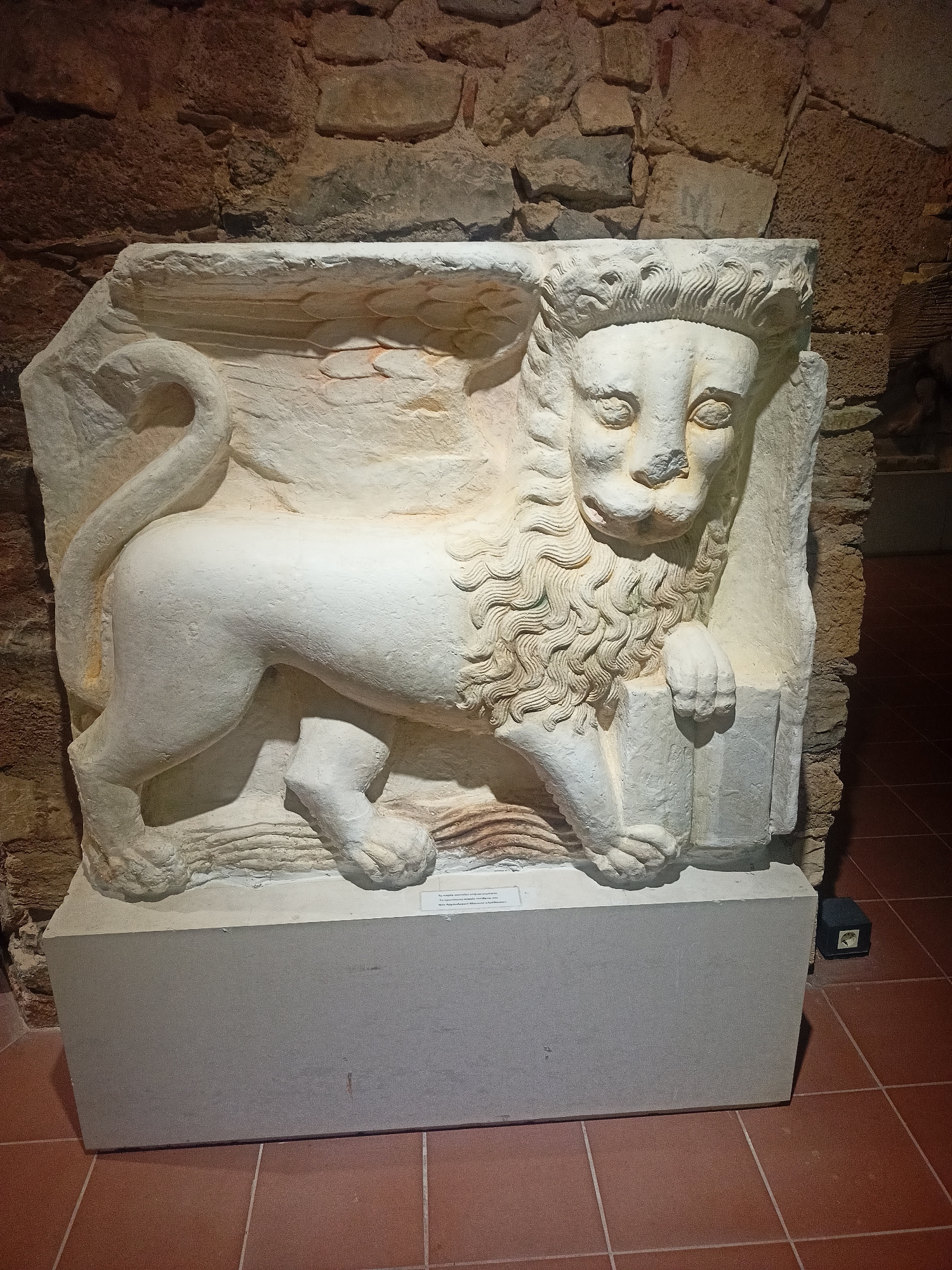 A stone relief of a winged lion is displayed on a light-colored rectangular pedestal against a rough stone wall in a museum in Chalkida, Greece. The lion, carved in white stone, is depicted in profile, facing left, with its body elongated and muscular. It has a mane of wavy curls, large expressive eyes, and a long, curled tail. Its front paws rest on an unseen object, and it appears to be standing on a wavy, textured surface. The background wall is composed of irregularly shaped, rough-hewn stones in varying shades of brown and gray. The floor is tiled with reddish-brown square tiles. A small black electrical box is visible on the floor to the right of the pedestal. A label is affixed to the front of the pedestal, though its text is not legible.