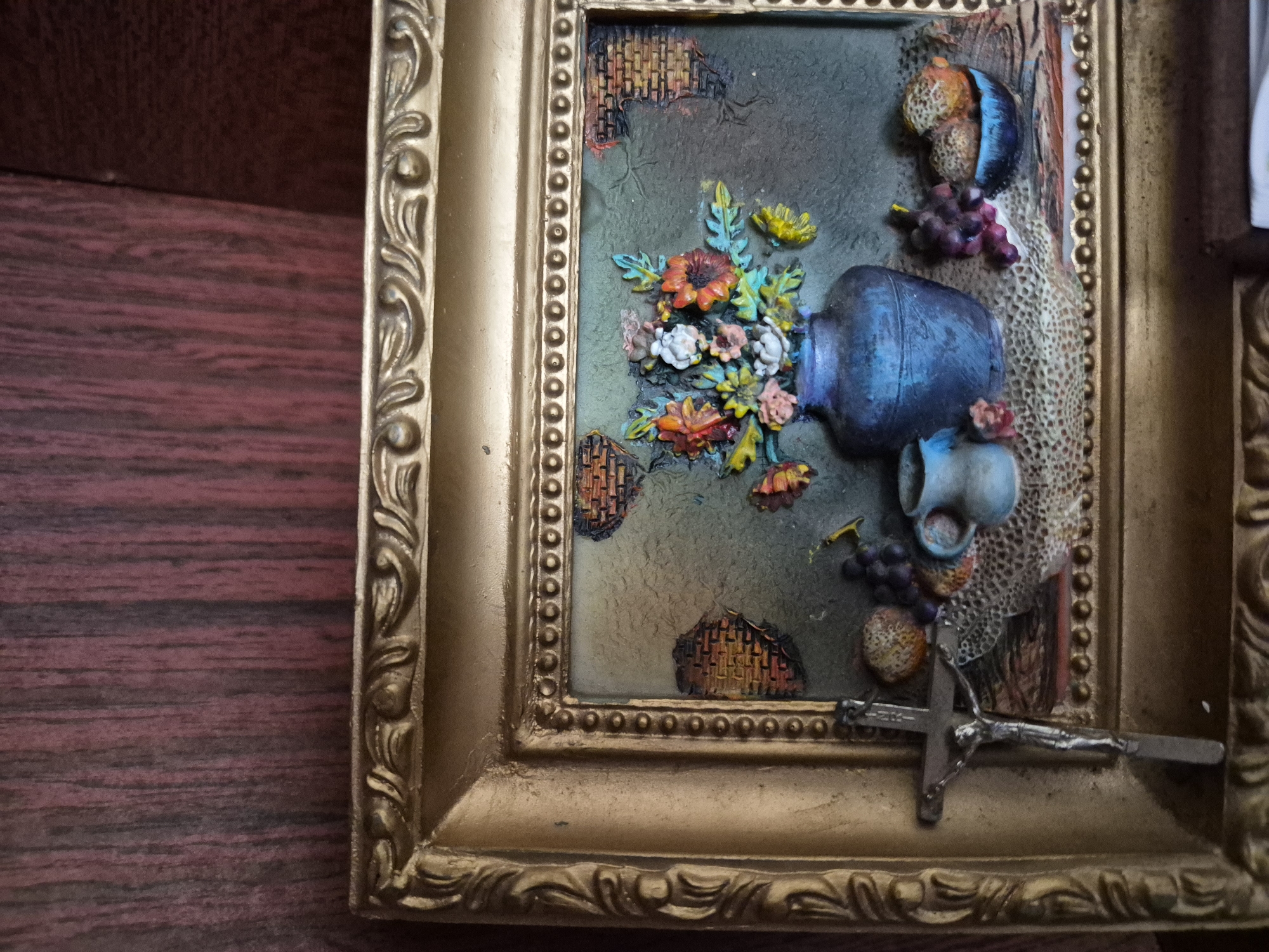 The image shows a framed still life artwork. The artwork depicts a blue vase filled with colorful flowers, a small light blue jug, a bunch of grapes, and a bowl of fruit. In the foreground, to the left of the artwork, is a silver crucifix hanging from the frame. The artwork is framed in a decorative gold-colored frame. The background of the image is a dark wood surface. The location in the city of Vanadzor, Armenia, is not specified in the image.