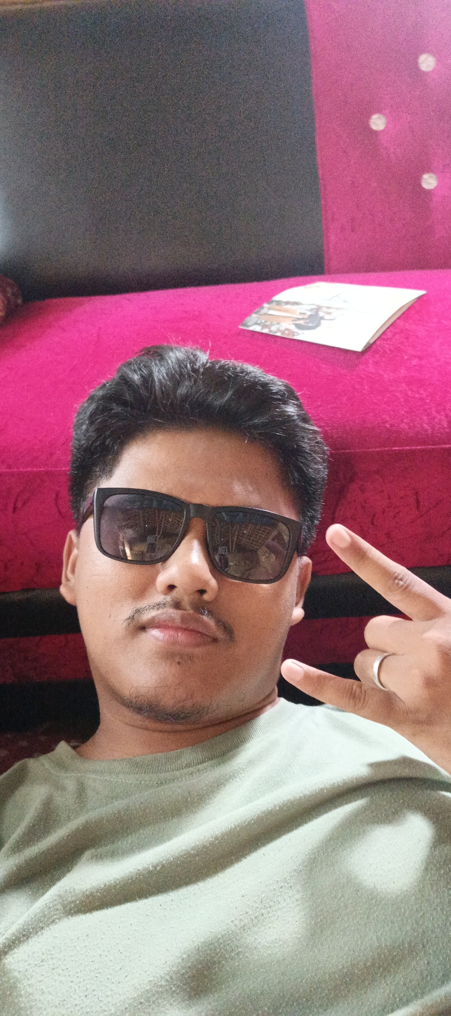 A young man wearing sunglasses and a light green t-shirt is pictured from the chest up, posing for a selfie. He has dark hair and a mustache. His right hand is raised, forming a peace sign with his index and middle fingers, while his left hand is partially visible. A silver ring is on his left ring finger. He is positioned in front of a vibrant pink surface, which appears to be a sofa. To the right of the man, a small, open booklet with visible illustrations is placed on the pink surface. The background consists of a dark, textured surface above the pink one. The lighting suggests an indoor setting. The overall impression is a casual, personal photograph taken in a home environment.