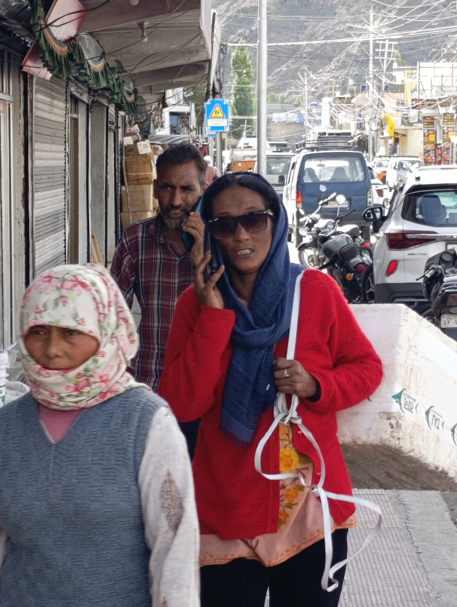 In Leh, India, a man in a plaid shirt talks on a phone while walking on a sidewalk. To his right, a woman in a red sweater and blue scarf, wearing sunglasses, also holds a phone to her ear. In the foreground, another woman wears a floral headscarf and a grey vest over a light pink top. Behind them, a street scene unfolds with various vehicles, including cars, motorcycles, and a blue van, along with storefronts and overhead power lines. The environment suggests a busy urban street with buildings lining one side and traffic on the road.