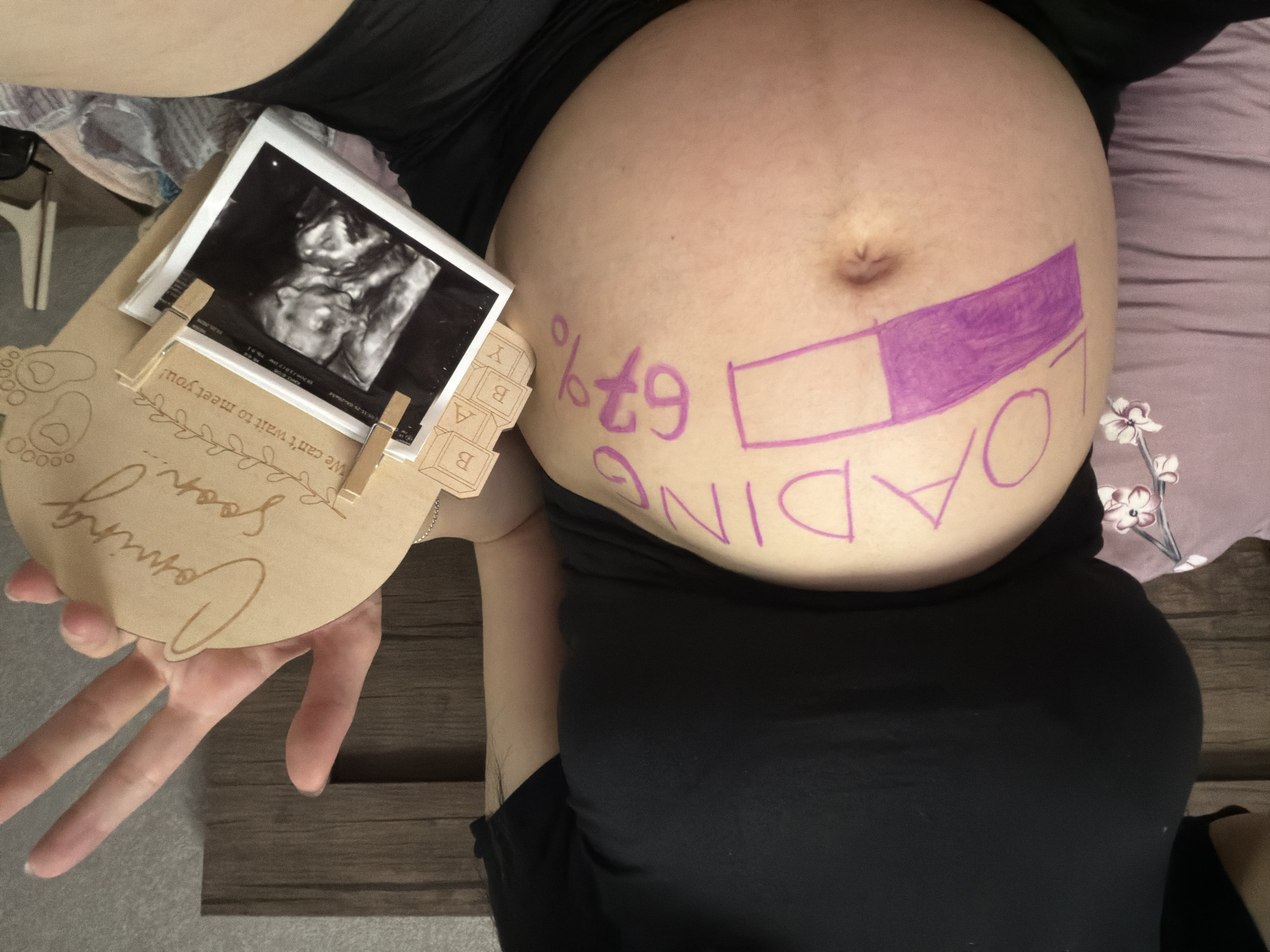 A pregnant person is lying on their back, with their belly facing upwards. They are wearing black clothing. On their belly, the word "LOADING" is written in purple marker, along with a progress bar filled to 67%. To the left of the belly, there are two ultrasound photos and a wooden plaque with "Coming Soon!" and "We can't wait to meet you!" engraved on it. The scene appears to be indoors, possibly a bedroom or living room, with a wooden floor and a patterned blanket visible. The lighting suggests it could be daytime. The overall mood is one of anticipation and excitement for the upcoming arrival of a baby. The location is identified as Tbilisi, Georgia.