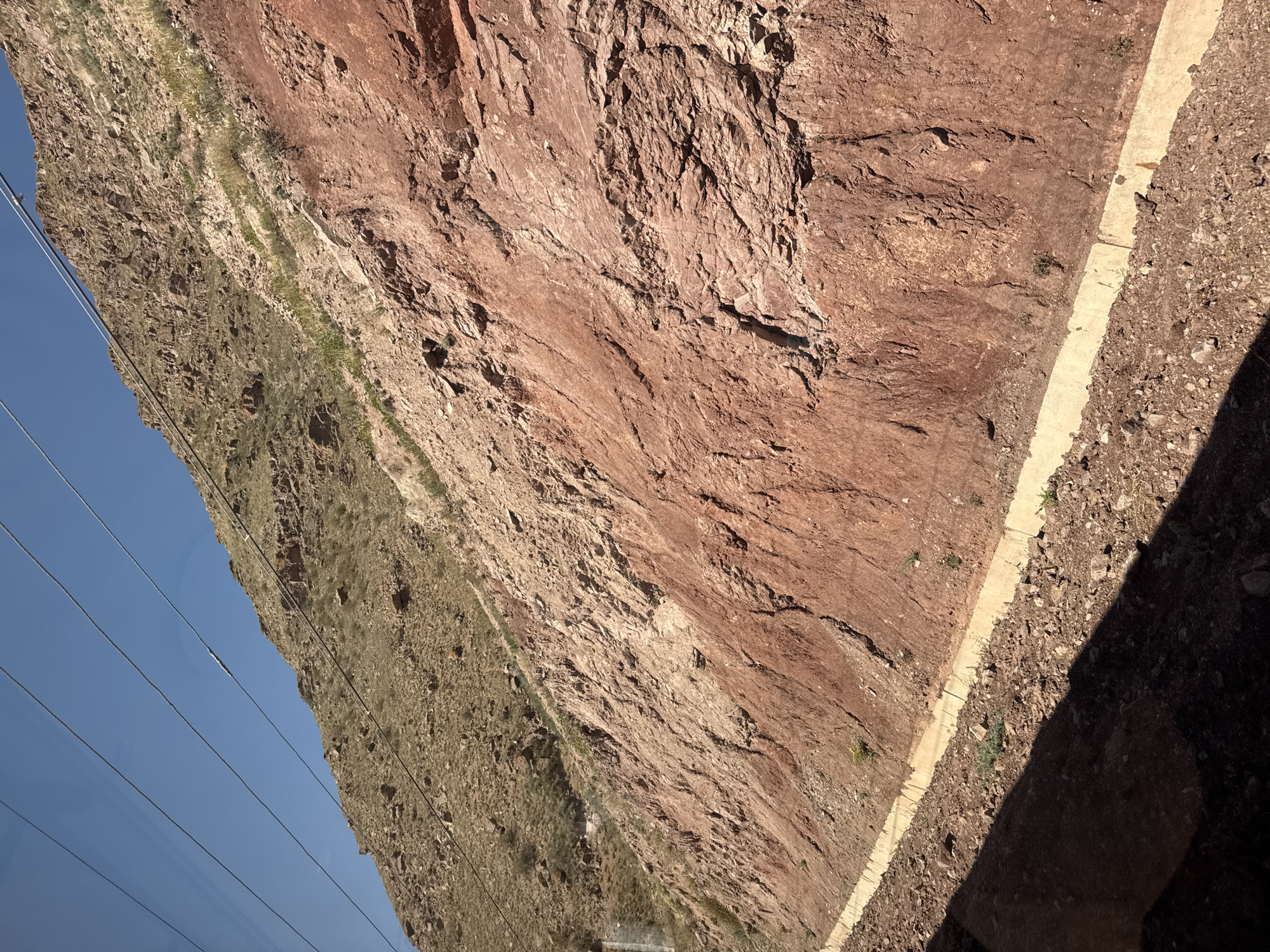 The image depicts a steep, rocky mountainside with exposed red rock layers and a patch of dry, brown vegetation on a higher slope. In the foreground, a concrete drainage ditch runs diagonally along the base of the mountainside, bordered by loose gravel and some sparse greenery. Overhead, several power lines stretch across the clear blue sky. The scene suggests a natural landscape with some human intervention for infrastructure. The location is specified as Krasnyy Most, Kyrgyzstan.