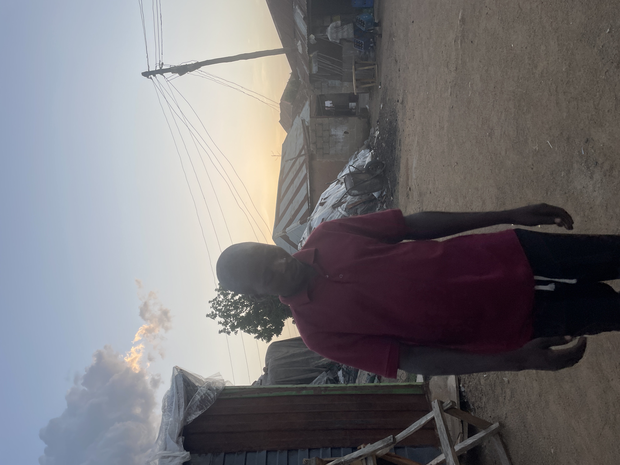 The image shows a man in Jos, Nigeria, wearing a red shirt and black pants. He stands on a dirt road in a residential area. Behind him are structures of varying materials, including metal sheeting and concrete blocks. Power lines are visible against a pale sky with clouds, suggesting either sunrise or sunset.