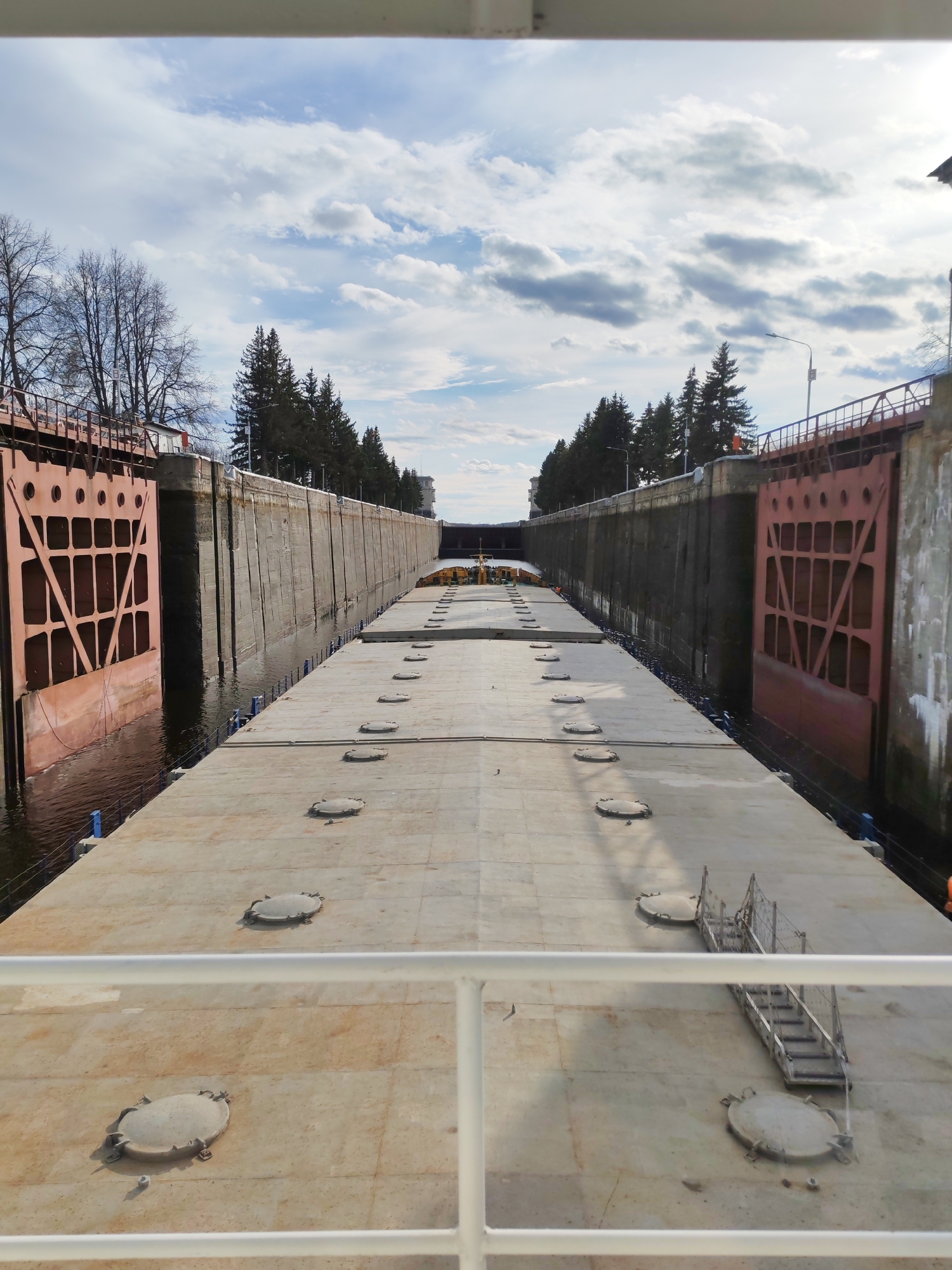 The image shows a view of a canal lock with large metal doors on either side. The lock is filled with water, and a large flat structure, possibly a barge or pontoon, occupies the center of the lock. The structure has several round hatches or access points on its surface.
On the sides of the lock, there are concrete walls with ladders and walkways. Trees can be seen growing along the banks of the canal.
The sky is partly cloudy. A white railing is visible in the foreground, suggesting the photograph was taken from a vessel or platform.
