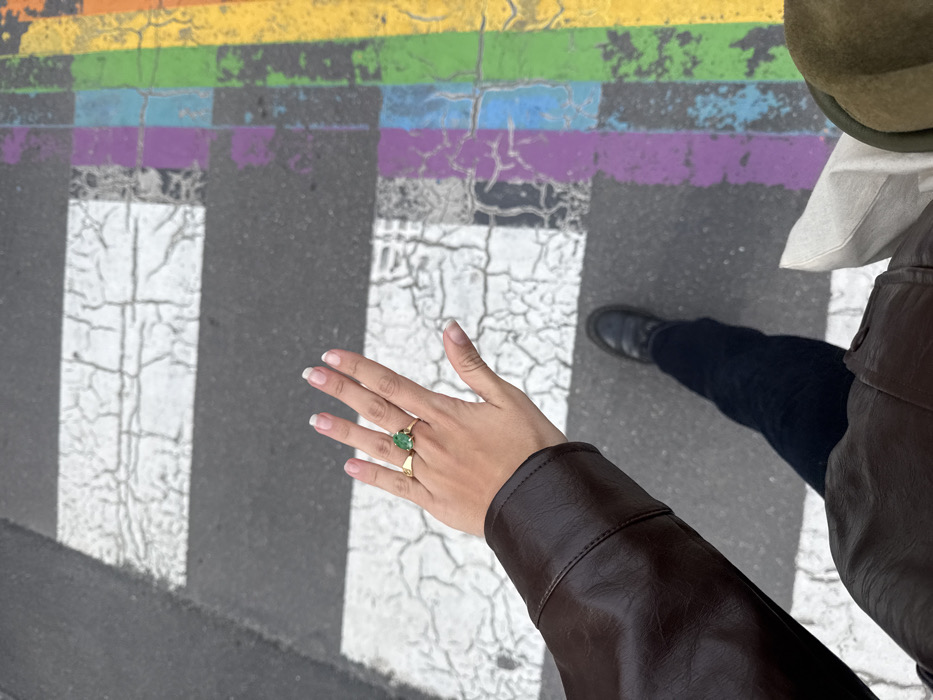 A high-angle, top-down photograph captures a person standing on a pedestrian crosswalk located at the intersection of Rue des Archives and Rue de la Verrerie in the Marais district of Paris, France. The visible left hand of the individual is raised and features clear-polished nails. A gold-toned ring with a prominent pear-shaped green gemstone is worn on the left ring finger, and a plain gold-toned band is on the left index finger. The individual wears a brown leather jacket, dark blue trousers, and a black chunky-soled loafer on their left foot. A light-colored bag and a section of a dark olive green hat are visible on the right side of the frame. The asphalt road beneath shows a standard pedestrian crossing with faded, cracked white stripes. Adjacent to these, a section of the crosswalk is painted with worn and chipped rainbow colors, indicative of the pride flag, displaying shades of purple, blue, green, yellow, and orange.