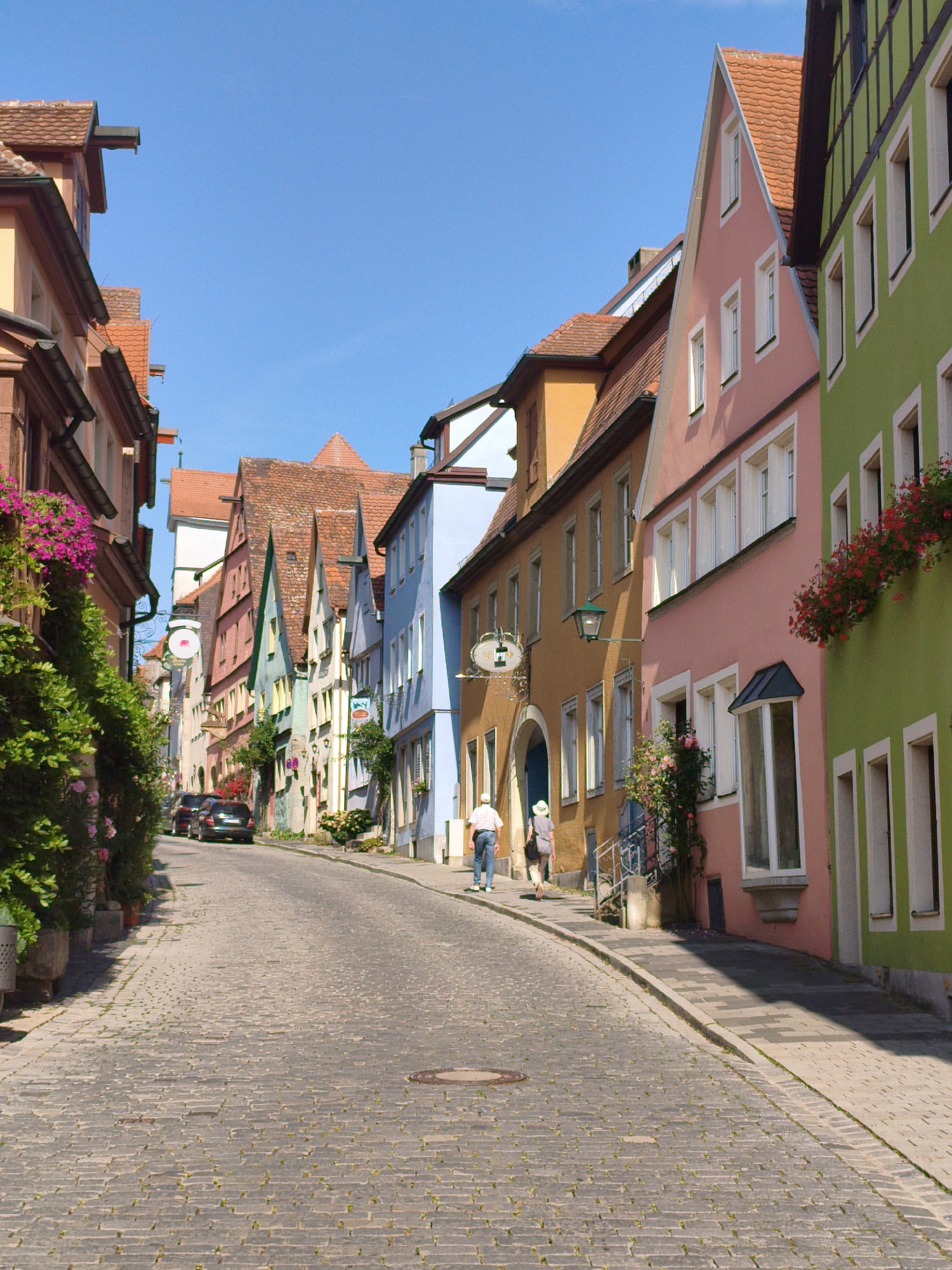 A cobblestone street winds uphill in Rothenburg ob der Tauber, Germany, lined with brightly colored, historic half-timbered buildings. The facades of the houses are painted in various pastel hues, including pink, green, orange, and blue, with traditional red-tiled roofs. Lush green foliage and pink flowers cascade from window boxes and planters, adding a vibrant touch to the scene.
Two individuals are walking up the street. The person in front is wearing a light-colored hat and a grey top with dark pants. The person behind them is wearing a white shirt and blue jeans. A dark-colored car is parked on the left side of the street. The sky is clear and blue.