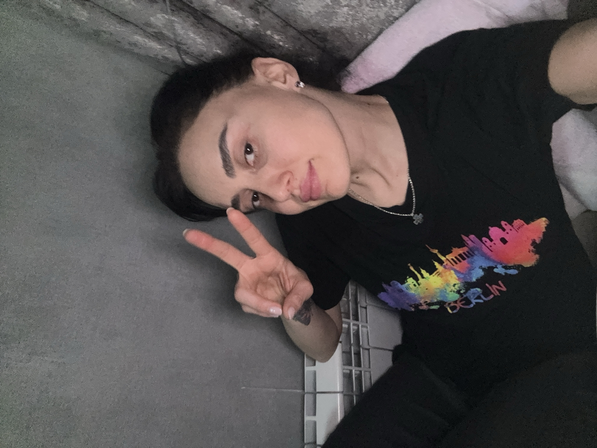 A young woman is lying on a gray floor or surface with her head resting on a light pink pillow or cushioned area. She is wearing a black t-shirt with a colorful print of the Berlin skyline and the word "BERLIN" beneath it. She is making a peace sign with her left hand. The background includes a gray wall and a white tiled area. The setting is ambiguous, but the presence of tiling suggests it might be a bathroom or similar space.