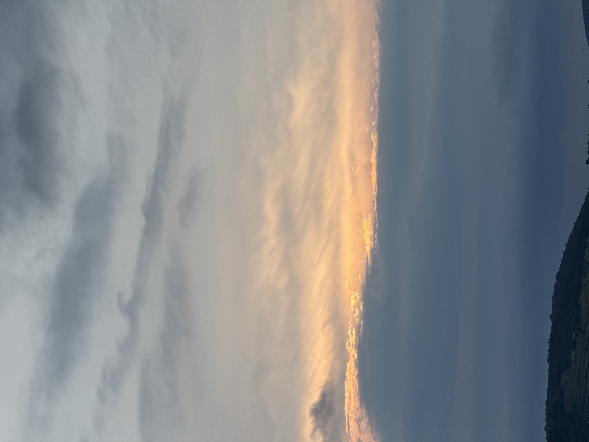 The image depicts a cloudy sky over the outskirts of Berezhany, Ukraine. A strip of golden light, indicating sunset or sunrise, is visible between layers of clouds. The lower portion of the image shows a dark, silhouetted hillside with sparse vegetation, and a small, indistinct structure that could be a tower or antenna near the right edge of the frame. The sky is dominated by various cloud formations, ranging from wispy, light-colored clouds higher up to denser, darker clouds towards the horizon. The overall scene conveys a natural landscape at a time of diffused daylight.
