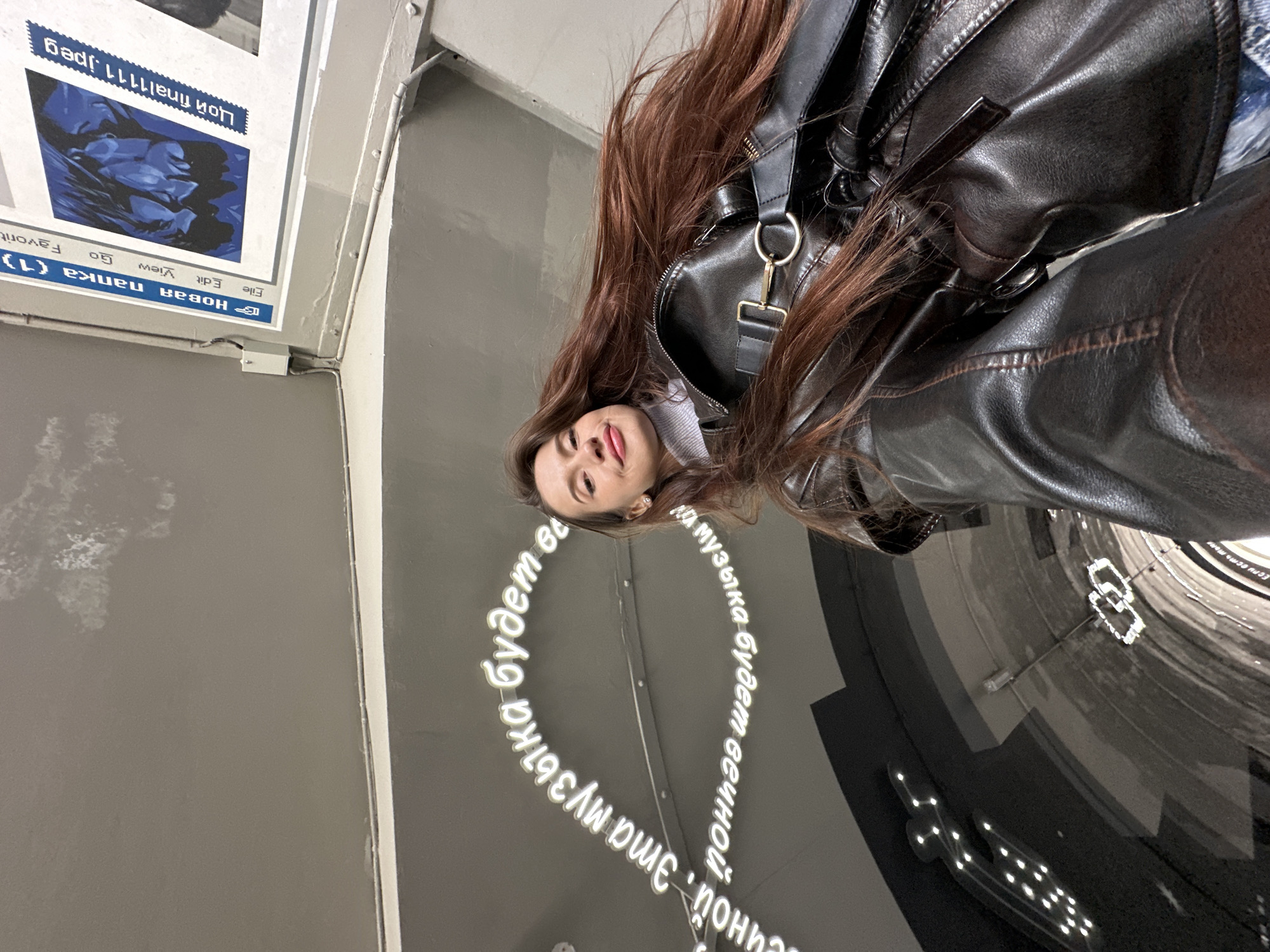A young woman with long brown hair is photographed indoors in Yekaterinburg, Russia. She wears a black leather jacket and a light gray turtleneck sweater. The camera angle looks up at her from a low position, creating a unique perspective.
She is standing next to a wall with a large, reflective, circular tunnel-shaped art installation. The phrase "Если есть вечной. Эта музыка будет вечной, Если есть вечной. Эта музыка будет вечной" in Russian is prominently displayed on the wall around the tunnel in white neon lettering. A sign with Russian words "Новая папка (1)" (New Folder (1)) and "Цой final1111.jpeg" can be seen in the upper-left corner of the image.