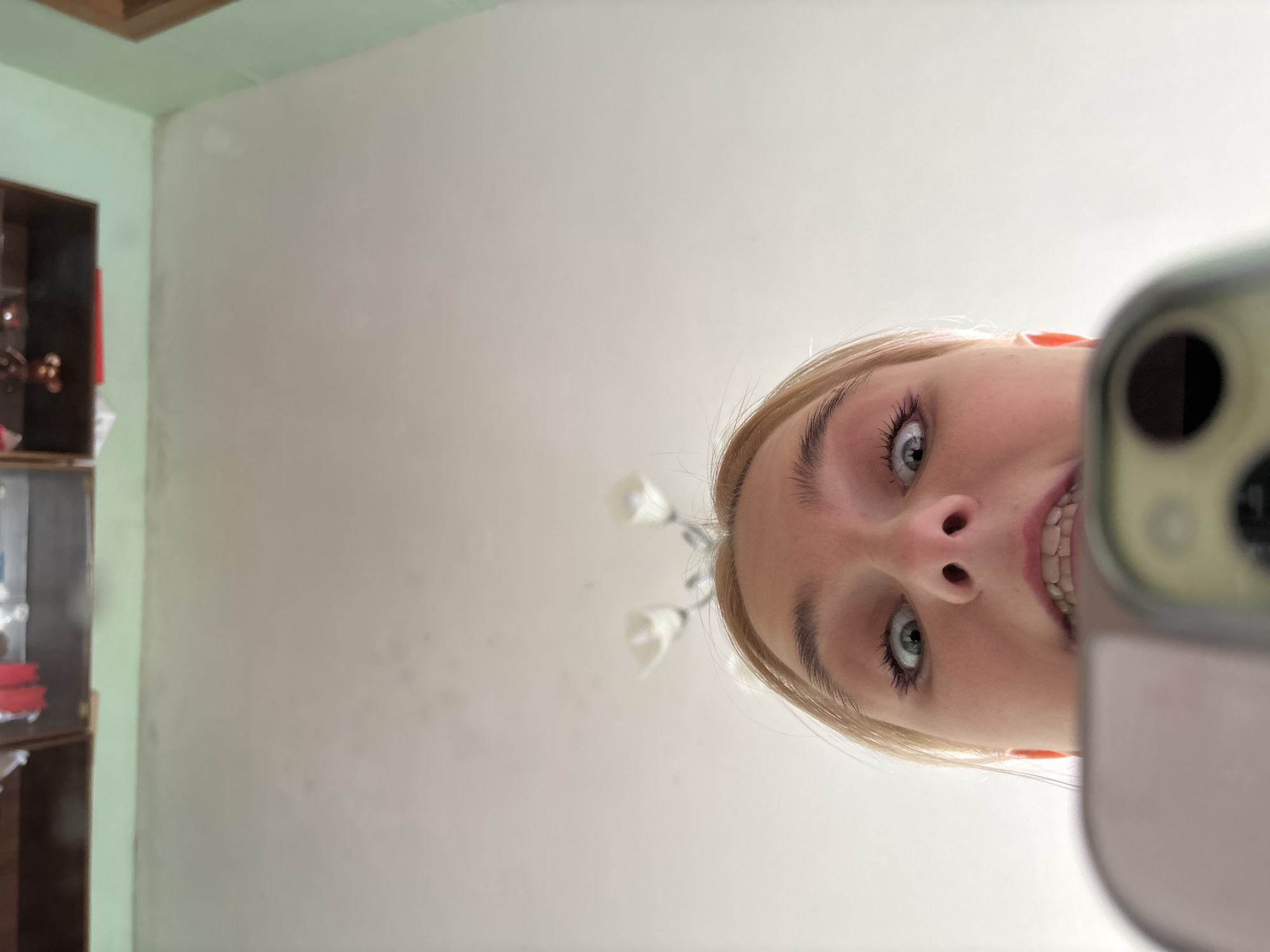 A low-angle, close-up, outdoor shot shows a young woman holding a smartphone. Her face is in focus, and she is looking directly at the camera with wide eyes and an open mouth, revealing her teeth. The phone is in the foreground, obscuring the bottom of her face. The background is out of focus, showing a wall and a ceiling with two lights. The lighting suggests it is daytime.