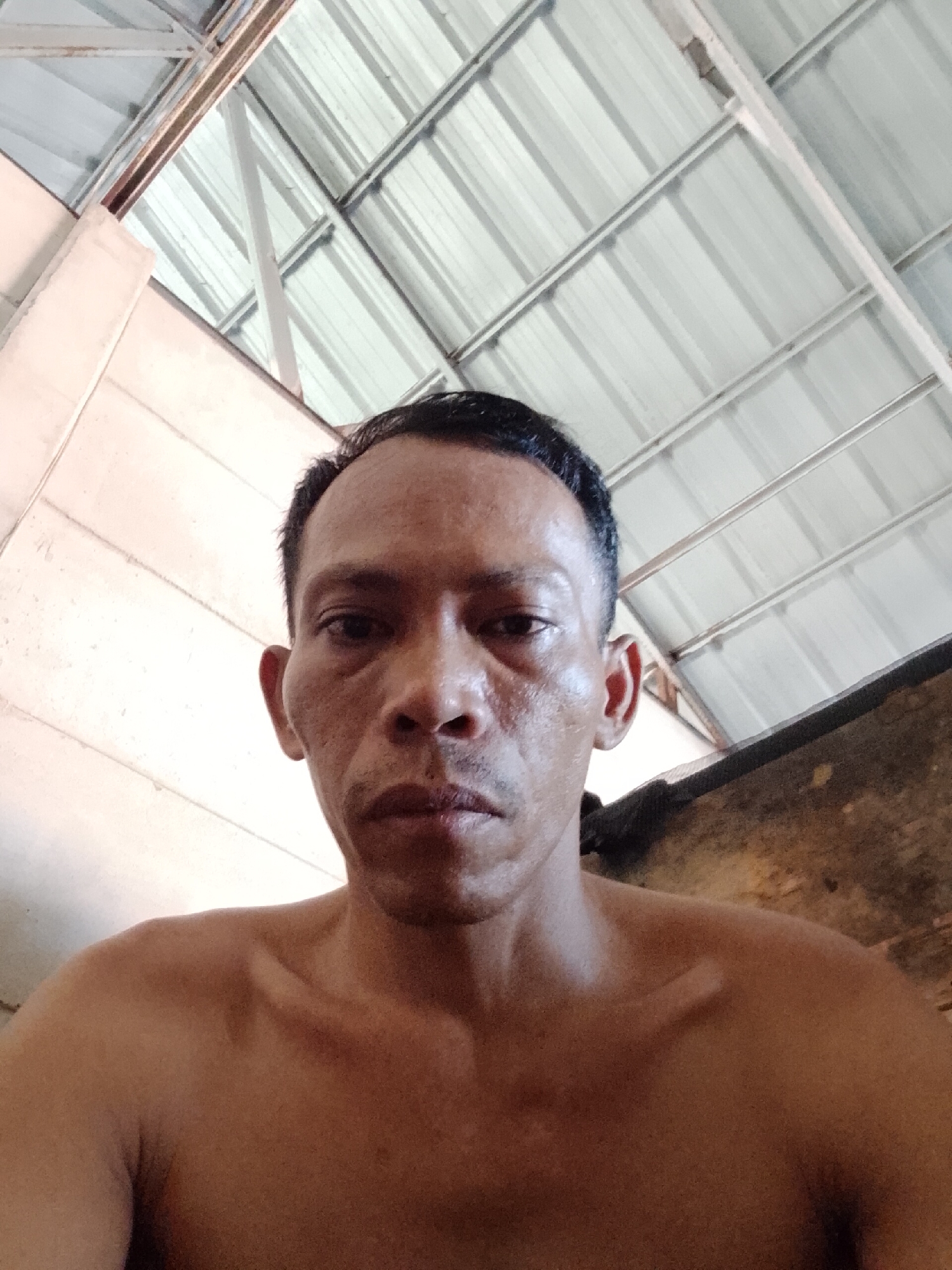 A man is visible from the chest up, gazing directly at the camera. He appears to be indoors under a corrugated metal roof supported by a white frame. The location is Bukit Ketok, Indonesia. The man's skin is brown. He has short, black hair and a serious expression. His upper body is bare. To the left is a flat wall. To the right is an unclear wooden structure or object. The lighting suggests it is daytime.