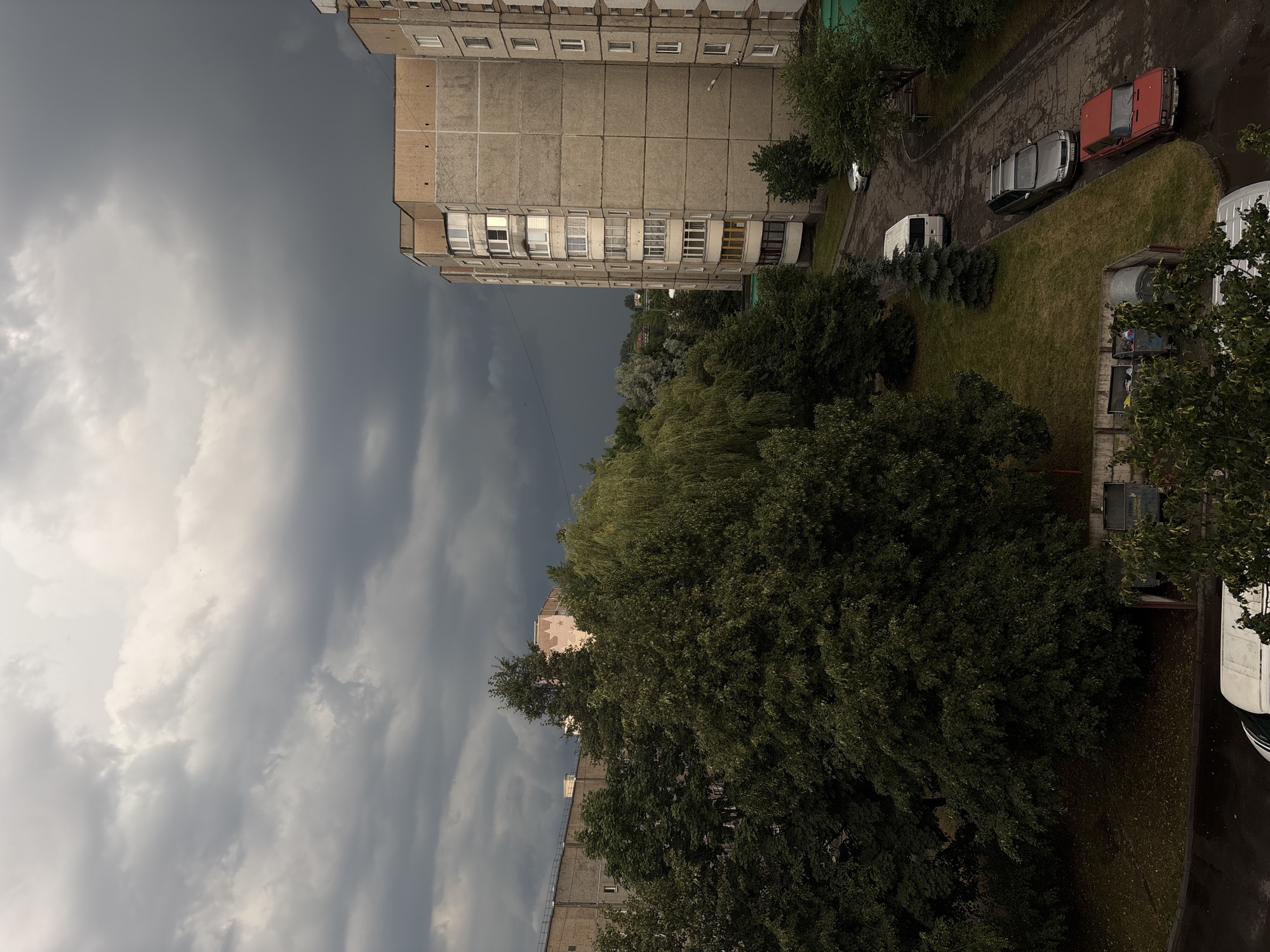 This is a vertical, outdoor, wide shot taken from a high vantage point, likely a building, overlooking a residential area. The scene is dominated by a dramatic sky filled with dark, heavy storm clouds. To the right, the perspective looks down on a grey, concrete apartment building with numerous windows, some of which are open.
Below and to the right of the apartment building, a patch of green grass is visible, interspersed with trees and bushes. A paved area or road runs alongside the grass, where several vehicles are parked. These include a red car, a silver car, and a white van. Further down the road, another white vehicle, possibly a van or minibus, is partially visible. A few more trees and parts of other buildings are also present in the background.
The time of day appears to be late afternoon or early evening, given the lighting and the stormy weather. The overall mood is somber and slightly oppressive due to the dark clouds, suggesting an imminent storm or the aftermath of rain. There are no visible people in the image. No text is discernible. The location is Lida, Belarus.