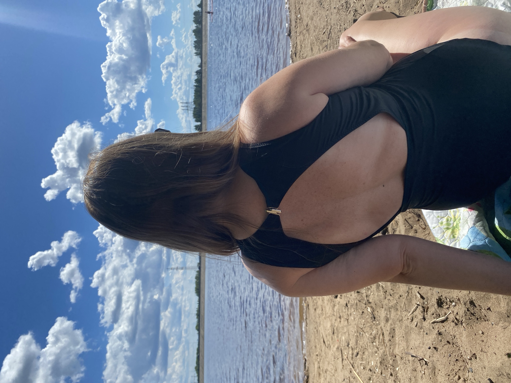 The image shows a woman from behind, lying on a sandy beach next to a body of water, likely a river or lake, in Noviy, Russia. She is wearing a black swimsuit with a cutout detail on the back. Her long, brown hair is flowing down her back. The sky is bright blue with fluffy white clouds, suggesting a sunny day. The water is calm and reflects the sky. The sandy shore is visible in the foreground, with some small pieces of driftwood scattered on it. In the background, a treeline is visible along the far bank of the water, with some tall industrial towers or pylons also present. A blanket with a floral pattern is partially visible beneath the woman. The overall impression is one of a relaxed, leisurely day at the beach. The time of day appears to be daytime, given the bright sunlight and blue sky.