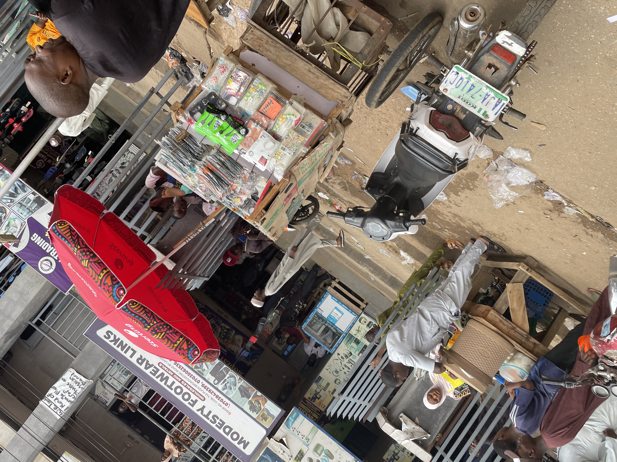 This image depicts a vibrant, bustling outdoor market scene in Maiburiji, Nigeria, during what appears to be daytime. The area is filled with people, market stalls, and a parked motorcycle, suggesting a busy commercial street.
Several individuals are present, engaging in various market activities. In the foreground, a man in a black shirt looks down, while another man, dressed in a white tunic and light trousers, carries a cardboard box. Nearby, a woman in a yellow top and headscarf stands. Other figures, partially visible in the background, contribute to the lively atmosphere.
Market stalls display a variety of goods. One wooden cart holds numerous small items, likely phone accessories such as cases and chargers, with visible branding like "oralmo," "2A," and "Hello," alongside the text "PHONES & ACCESSORIES." Another display area advertises "MODESTY FOOTWEAR LINKS," offering "WHOLESALES AND RETAILS" with phone numbers "07038429399" and "07035062536," and a location "QSHOP NO, CG08, 740 PLAZA BROADCASTING ROAD BY LAGOS."
A prominent red umbrella, adorned with patterns and the word "Sterling," provides shade over one of the stalls. A white and black scooter/motorcycle is parked, displaying a Nigerian license plate from Kogi State: "JJA-741QC." The ground shows some litter, including plastic bags, typical of a busy street environment.
Other visible texts include "TRADING," "K.W TRADI," "MANDII," and "ALIYUR." A handwritten sign reads, "We ben Kind of crap Anage Plic Phone an siem Panel," possibly indicating services for phone screen or panel repairs/sales. The scene captures the dynamic daily life of a Nigerian street market.