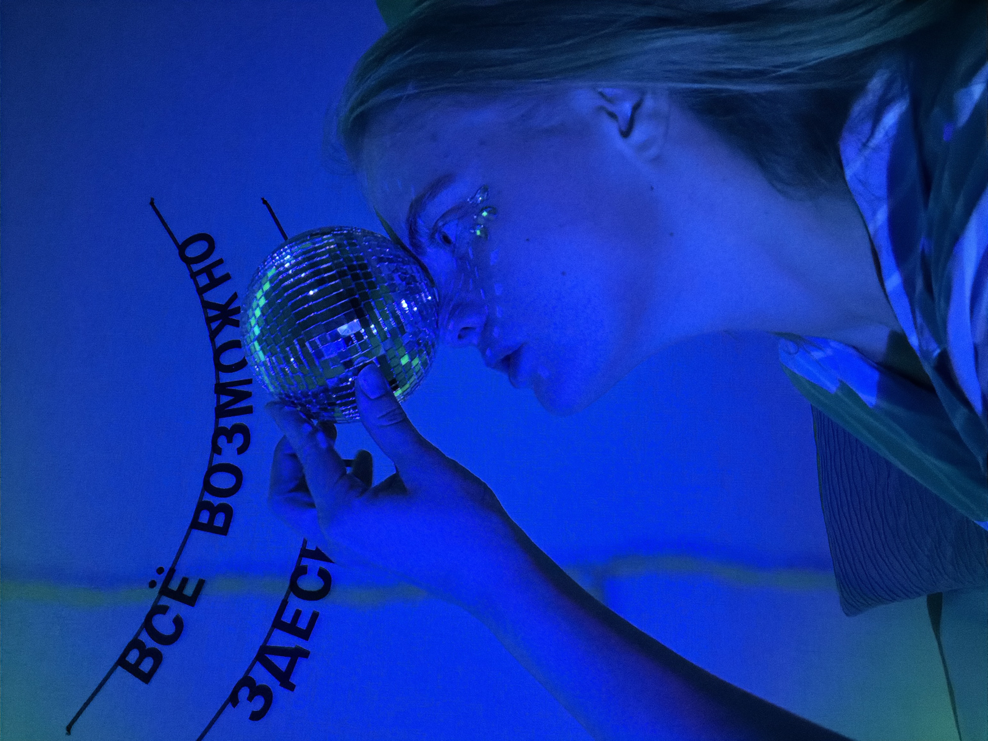 A woman with blonde hair and glittering makeup near her eye is holding a disco ball in her left hand and tilting it toward her face. She is wearing a patterned top or robe. To the left, in the background, are Russian words. The blue lighting makes identifying the location in Izhevsk, Russia, difficult.