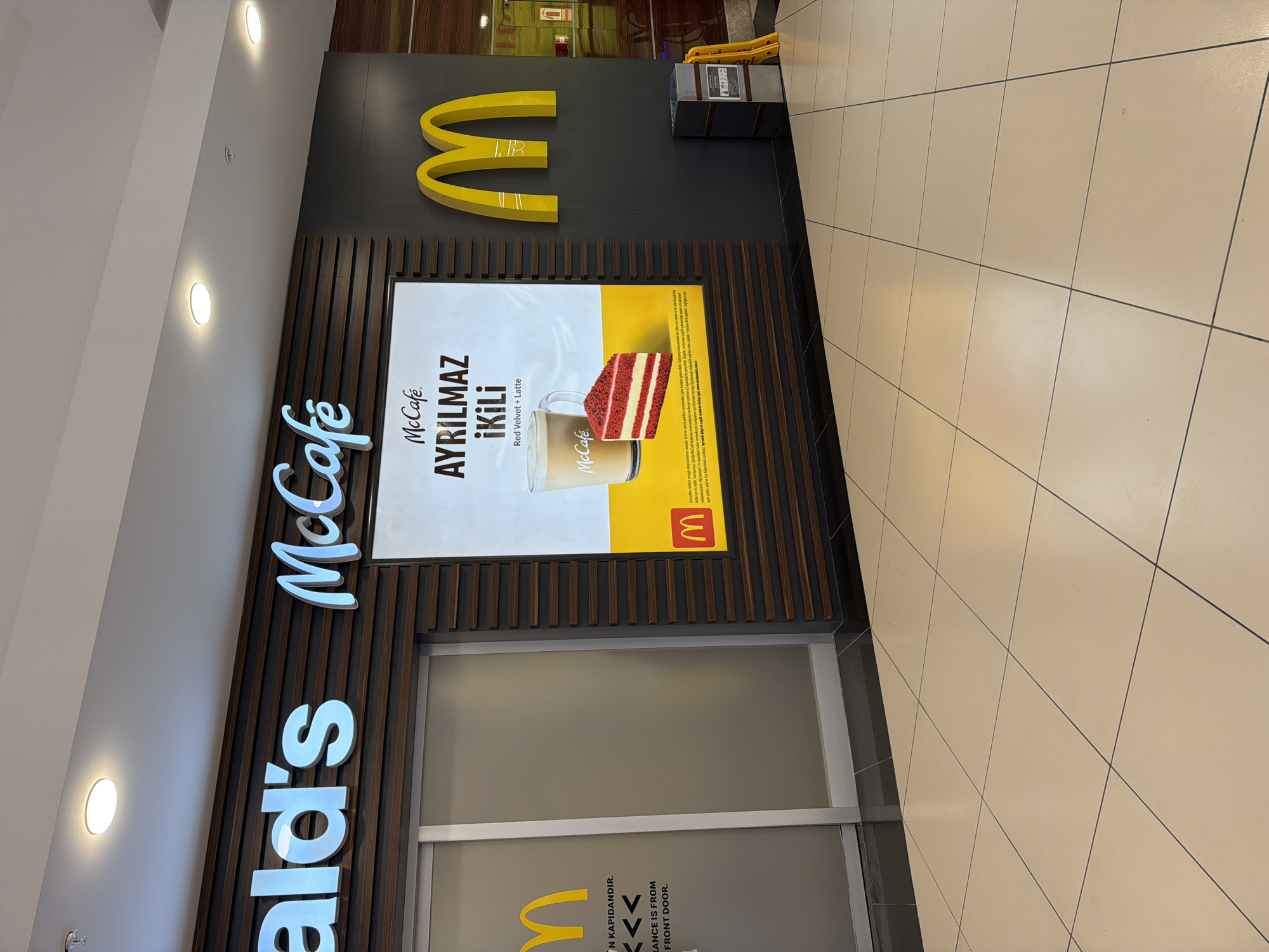 The image displays the storefront of a McDonald's McCafé, likely situated within an indoor commercial building such as a shopping mall in Alanya, Turkey. The scene is brightly lit by overhead ceiling lights, indicating an indoor environment.
The main subject is the McCafé entrance, featuring prominent signage. A large yellow McDonald's "M" logo is fixed on a dark grey wall, alongside the glowing white and blue "McCafé" and partially visible "McDonald's" signs. Below the "McCafé" sign, wooden vertical slats form part of the facade.
A large promotional poster is centrally displayed, advertising "McCafé AYRILMAZ İKİLİ" (meaning "Inseparable Duo"), featuring a "Red Velvet + Latte." The poster shows a slice of red velvet cake next to a glass of latte, with the McCafé logo prominently displayed on both the glass and the poster. Smaller Turkish text on the poster likely details promotional terms.
Below the poster is a frosted glass entrance door. Text on the door reads "GİRİŞ KAPI'DANDIR." and "ENTRANCE IS FROM FRONT DOOR.", indicating the entry point. To the right of the "M" logo, there's a grey utility unit or trash bin, with yellow safety barriers or cones nearby. The floor is covered with light-colored square tiles, typical of commercial interiors. No people are visible, and no specific activity is taking place other than the static display of the storefront. The time of day is not discernible due to the indoor setting and artificial lighting.