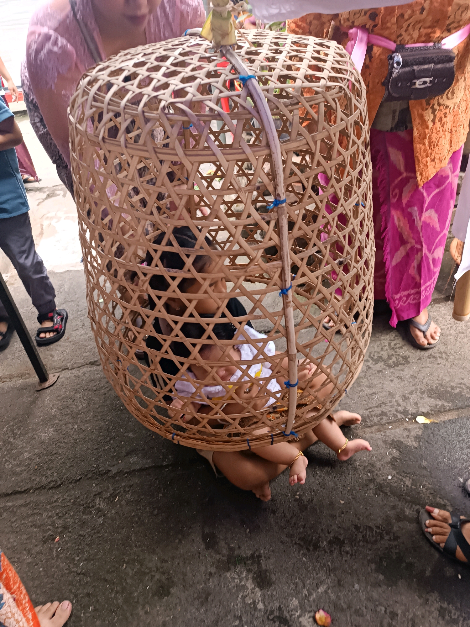 In Tabanan, Indonesia, a young child is positioned inside a large, woven bamboo basket, which is elevated from the ground. The child appears to be dressed in white clothing. Several adults are present, with some of them holding the basket, suggesting they are part of a procession or cultural event. One adult to the left wears a blue shirt and grey pants, while another to the right is adorned in traditional orange and pink patterned attire. The ground appears to be paved, and the overall setting suggests an outdoor community gathering.