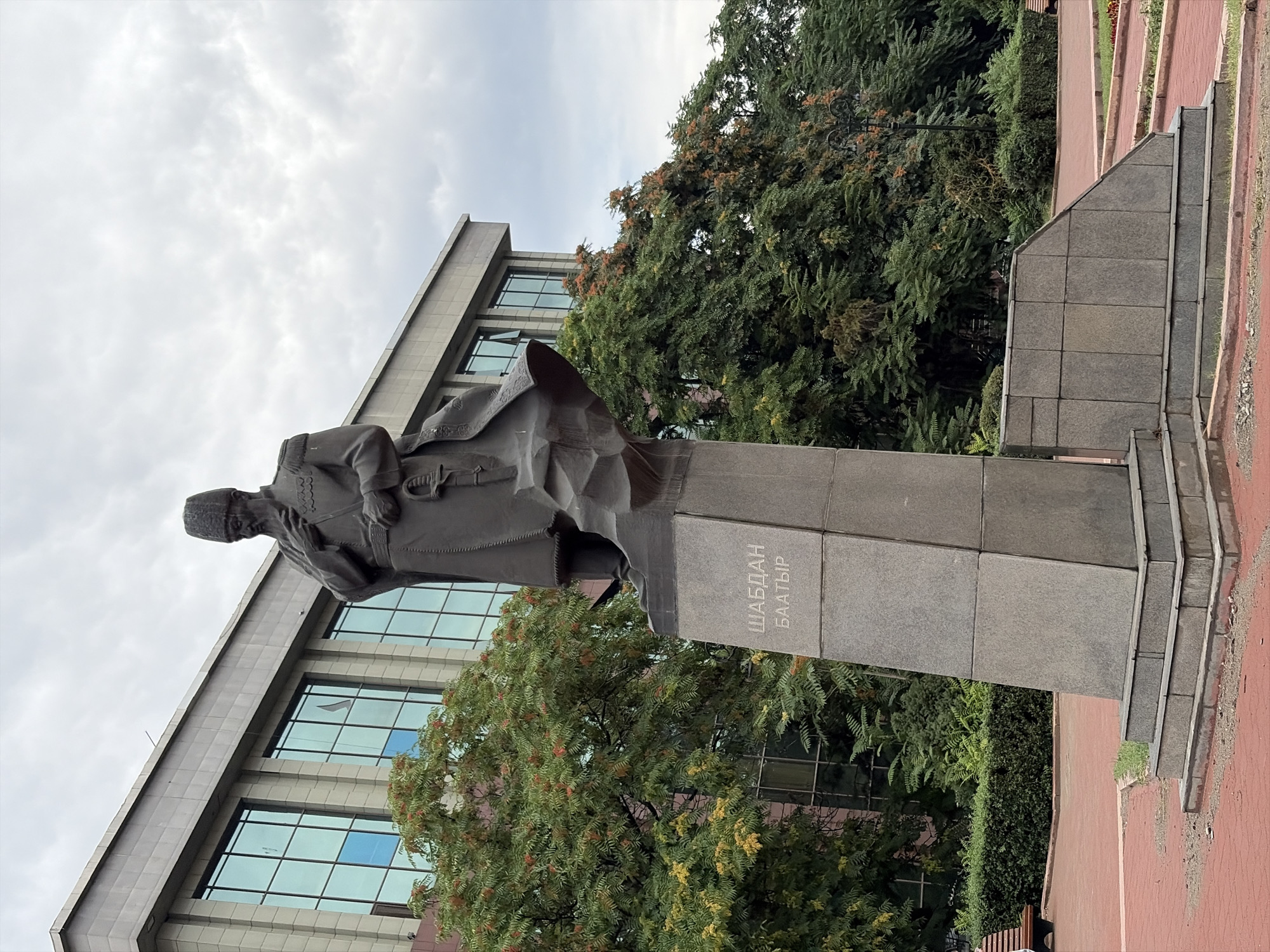 A statue of a man in traditional clothing, possibly military attire, stands on a large stone pedestal in a park. The man has a beard and is wearing a hat. The statue is dark in color, likely bronze or stone. The pedestal has text inscribed on it, which reads "ШАБДАН БАТЫР". In the background, a modern building with large glass windows is visible. Trees with green and orange foliage surround the statue and the building. The ground in the foreground is paved with red bricks. The sky is overcast with clouds. This monument is located in Bishkek, Kyrgyzstan, and is dedicated to Shabdan Batyr, a Kyrgyz warrior and statesman.