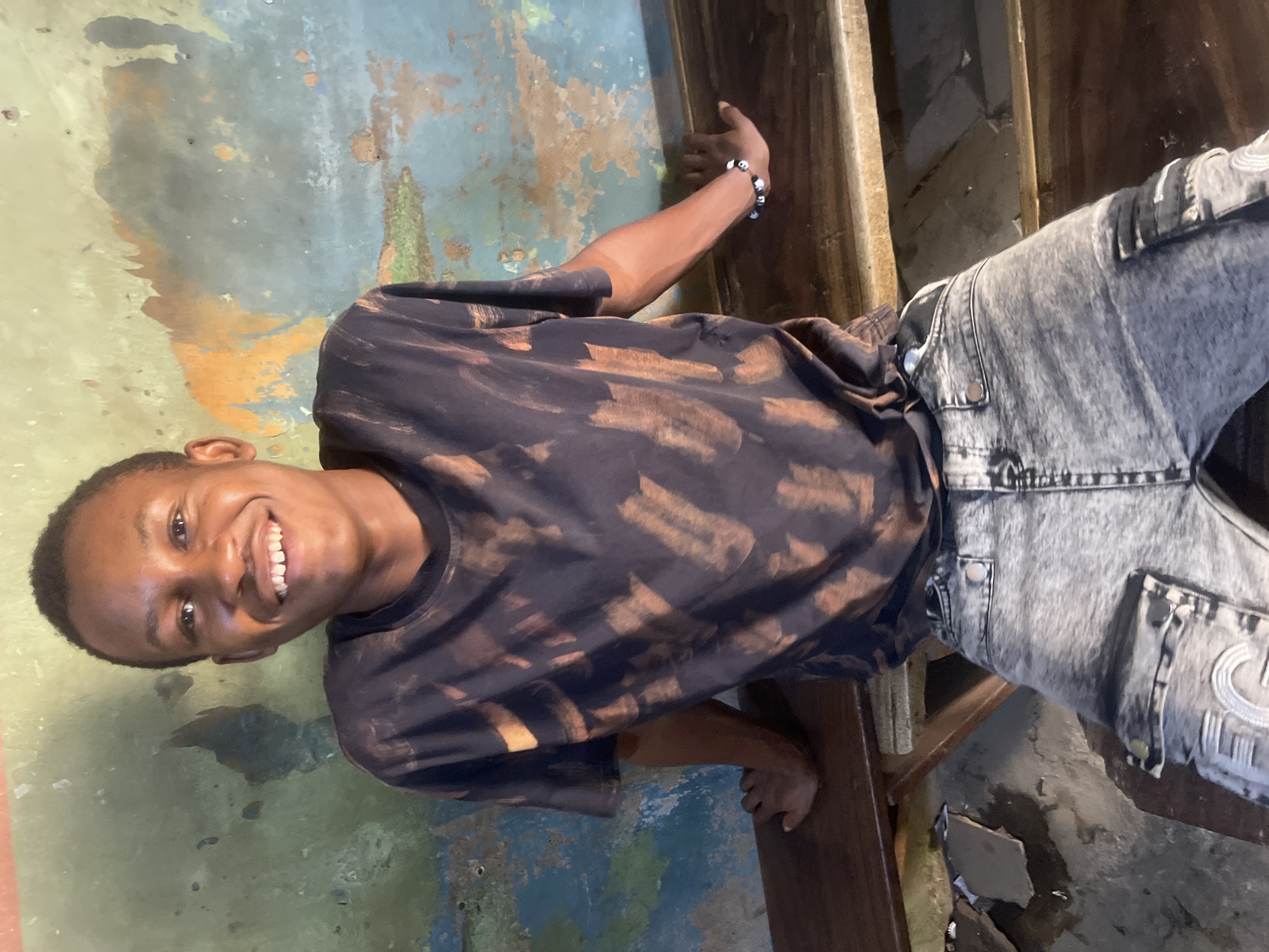 A smiling young man stands in what appears to be an indoor setting. He wears a dark t-shirt with bleached patterns and faded gray jeans. The background features a distressed wall with peeling paint in shades of blue, green, and orange. Adjacent to him is a wooden structure. The presence of distressed walls and the wooden structure may suggest an older, possibly informal, setting. While the image is taken in Zaria, Nigeria, there are no further details visible to provide more specific information about the location in Zaria.
