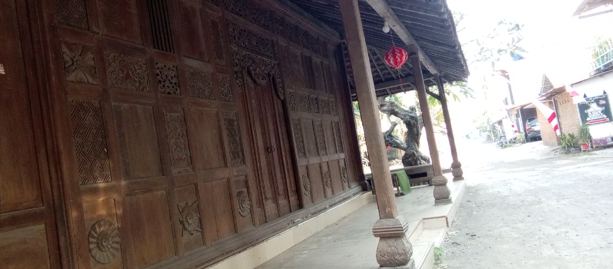 This image captures a detailed view of the intricately carved wooden facade of a building, possibly a traditional Javanese Joglo house, in Kediri City, Indonesia. The wood panels exhibit elaborate carvings of geometric patterns, floral motifs, and circular designs, showcasing skilled craftsmanship. Supported by thick, carved wooden pillars, the structure has a covered walkway or veranda running along its front. A red and white paper lantern hangs from the roof, adding a touch of festive decoration. Beyond the veranda, a gnarled, ancient tree is visible, its branches reaching out. The background reveals a glimpse of a street scene with a few buildings, a parked car, a motorcycle, and some potted plants. The overall impression is one of cultural heritage and a serene, traditional setting within an urban environment.