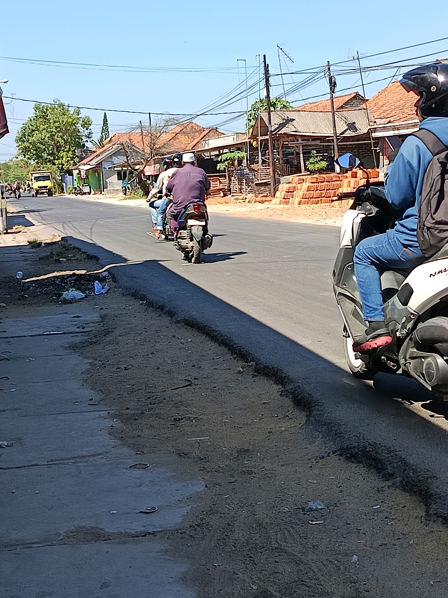 The image depicts a sunny day on a paved road in Blega, Indonesia. In the foreground, a person on a scooter, wearing a blue hoodie and a black helmet, is partially visible. They are riding on the right side of the road. Further down the road, three people are riding on two separate scooters. One scooter carries two individuals; the one in front wears a white cap and a dark purple shirt, and the one behind wears a light-colored shirt and blue jeans. The second scooter is ridden by a single person in a light-colored shirt and blue jeans. The background shows a line of houses with orange tiled roofs, some trees, and utility poles with numerous wires. A yellow truck is visible in the distance. The road appears to have some debris on the side, including discarded plastic bags and other waste. The sky is clear blue, suggesting a warm climate.