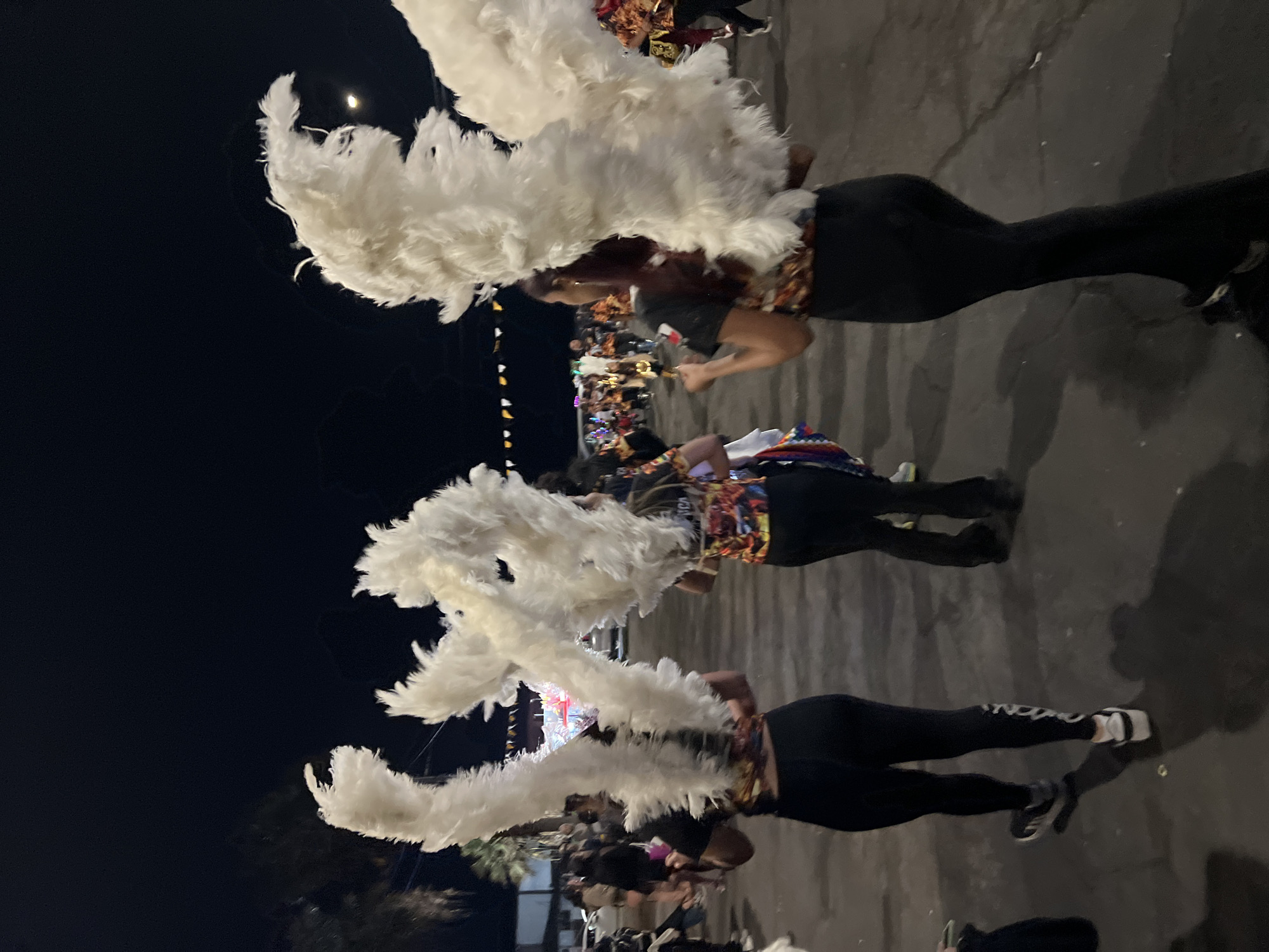 Three individuals are seen from behind, walking away from the camera at night on a paved street. They are all wearing black leggings and black t-shirts with colorful graphic prints on the back. Each person is wearing large, white feathered headdresses resembling wings, which are attached to their backs and extend upwards and outwards. The person on the left has white sneakers with grey accents, the middle person has black and white sneakers, and the person on the right has black shoes. In the background, other people and a few vehicles are visible, suggesting a street event or parade. Colorful flags or banners are strung across the street in the distance. The general scene appears to be outdoors in an urban environment at night. The image was taken in the city of Arica, Chile.