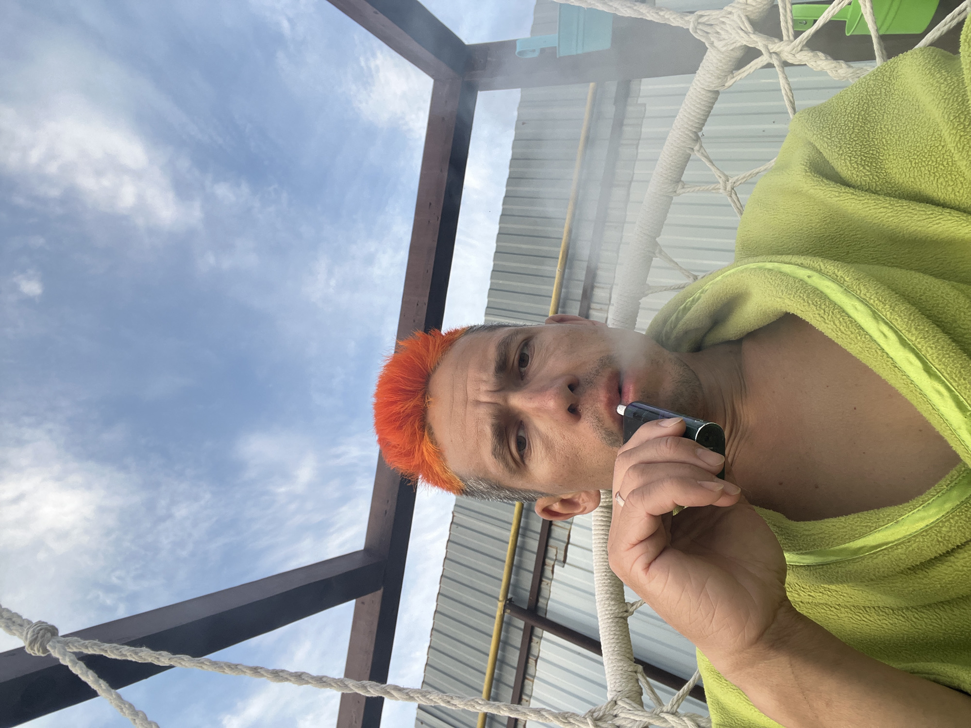 A man with orange hair is seated in a rope hammock. He is wearing a light green robe and smoking a vape pen. Smoke is visible in the air. The sky is visible in the background. Above the man are metal roof and wooden beams. The image appears to be taken outdoors, though specific information about the city of Chaikovsky, Russia is unavailable.