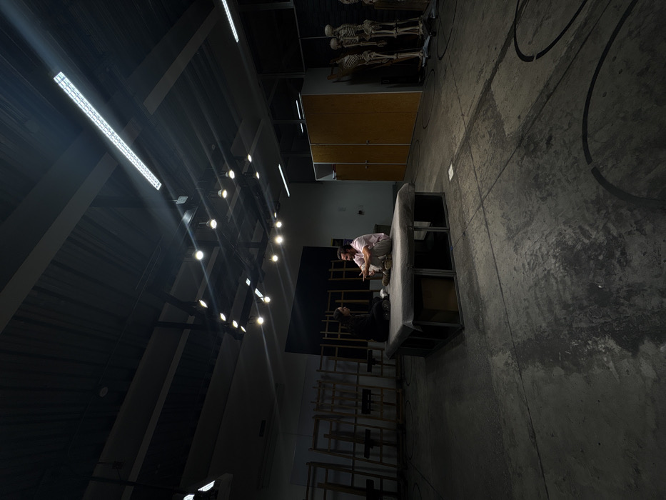 A person crouches on a platform, engaged in a conversation with another person sitting below. The backdrop features a dark wall and rows of wooden chairs. Fluorescent and globe lights illuminate the scene from above. To the right, there are storage cabinets and skeletal models. The floor has painted circles.