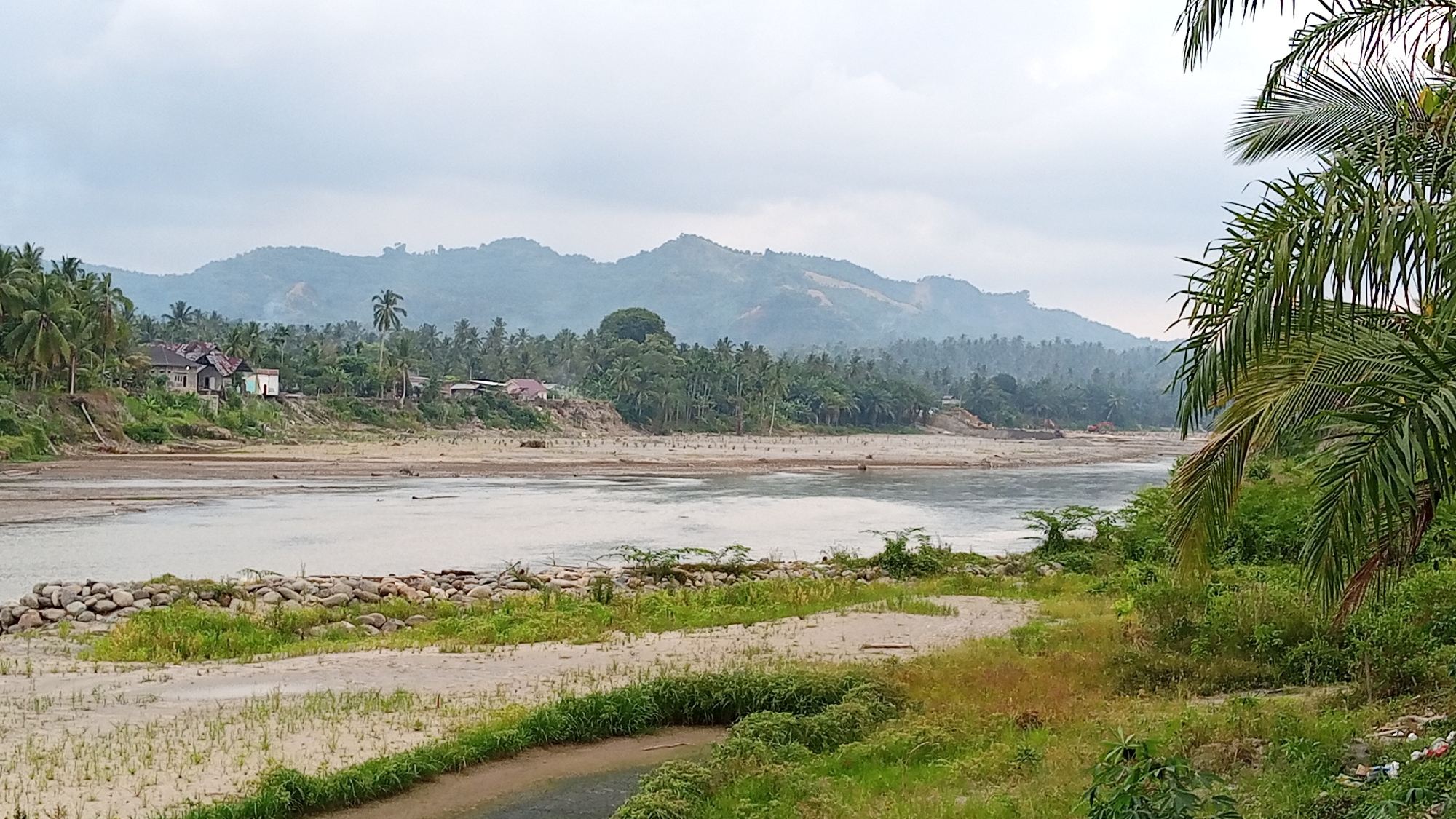 This image depicts a hazy, overcast day in Beunyot, Indonesia, with a wide river dominating the foreground. The river's banks are lined with sand and sparse vegetation, with a line of large rocks forming a barrier on the near bank. In the midground, a dense jungle of palm trees and other foliage covers the landscape, interspersed with a few buildings, including houses with corrugated metal roofs. A dirt road with patches of grass winds through the sandy area in the foreground. In the background, mist-shrouded mountains rise, their slopes covered in vegetation. The overall impression is one of a rural, tropical environment.