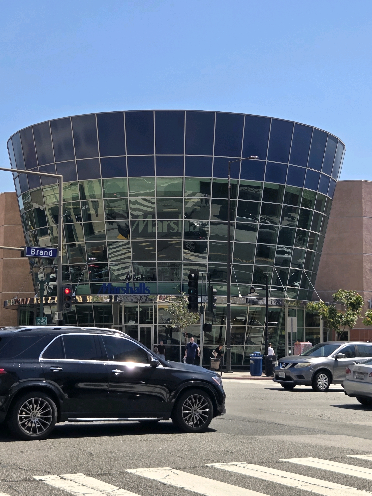 A medium shot captures a modern, curved glass-fronted building at the intersection of Brand Boulevard and an unnamed street in Glendale, California. The building, identified by a "Marshalls" sign visible through the glass, reflects the clear blue sky and surrounding elements.
In the foreground, a black SUV is parked on the right side of the frame, positioned at a crosswalk. To its right, a silver SUV is in motion, crossing the intersection. Behind the silver SUV, a portion of a light-colored vehicle is visible.
Several individuals are present near the entrance of the Marshalls store. One person, dressed in a dark blue shirt, stands outside. Another person, wearing a light-colored shirt, is seen near a blue trash receptacle. A woman in a dark top is also visible near the entrance, holding a bag.
A street sign reading "Brand" is mounted on a pole to the left of the building. A traffic light, currently showing red, is visible above the sign. The environment suggests a sunny day in a commercial district.