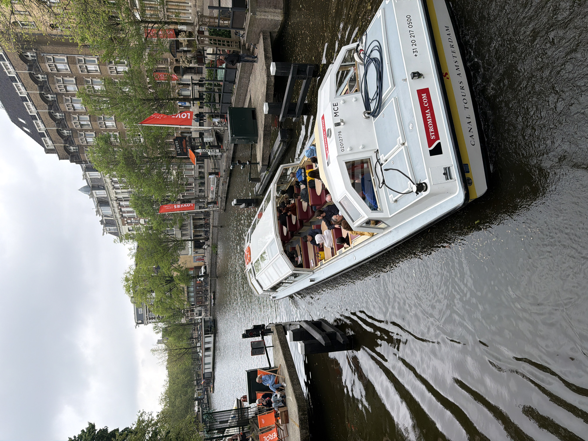 The image captures a canal tour boat, identified as "Stromma Canal Tours Amsterdam," navigating the canals of Amsterdam, Netherlands. The boat is white with red and yellow accents and carries passengers. The canal is lined with traditional Dutch buildings, with "Lovers Canal Cruises" signage and red flags visible along the shore. People are seated in orange chairs, likely part of a canal-side cafe or seating area. The water reflects the overcast sky.