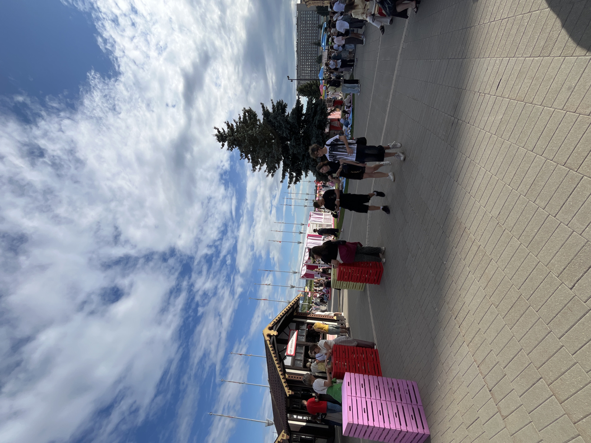 A sunny day unfolds in Nizhny Novgorod, Russia, where a lively outdoor market scene is captured. The foreground showcases a paved walkway, with neat rows of interlocking bricks creating a clean, urban pathway. To the left, stacks of vibrant pink and red crates are arranged, hinting at commerce and goods. In the mid-ground, a small wooden structure, possibly a food stall or vendor booth, is the center of activity. Several people are gathered around it, with one individual in a red shirt and another in a green apron attending to customers.
Further into the scene, the path opens up to a more expansive area populated by a diverse crowd. People are milling about, some walking, others sitting on the ground, suggesting a relaxed, festive atmosphere. A prominent, dark green pine tree stands tall in the middle distance, providing a natural focal point amidst the urban setting. Beyond the tree, a large, multi-story building with numerous windows dominates the background, indicative of the city's architecture. The sky above is a canvas of blue, adorned with scattered white clouds, casting dynamic shadows on the ground below. The overall impression is one of a bustling public space, likely a park or a designated event area, filled with people enjoying an outdoor gathering in Nizhny Novgorod.